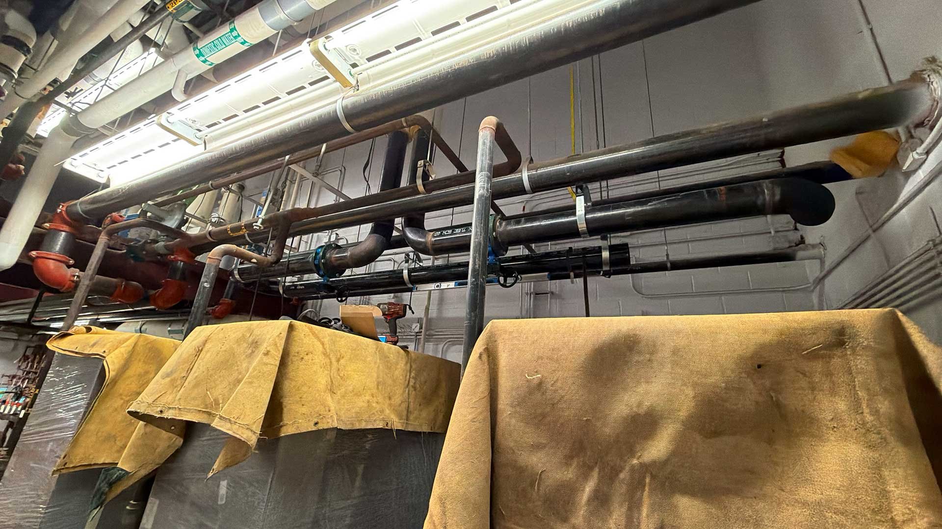 Industrial piping system with metal pipes, valves, and insulation, likely in a utility or processing plant. Covered equipment or containers in the foreground.