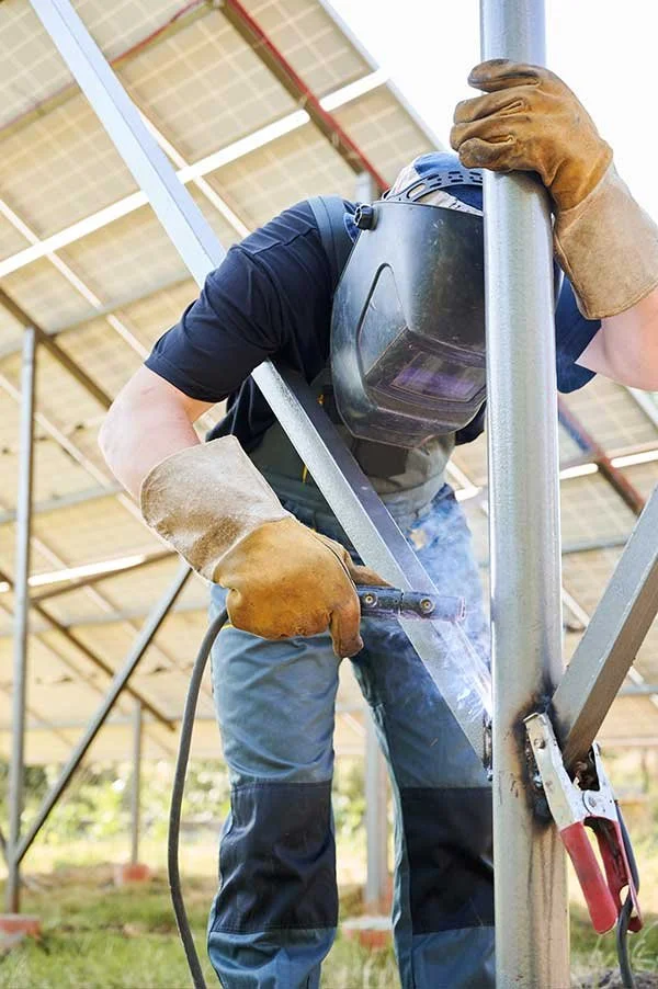 A person welding metal at an outdoor workshop, wearing safety gloves and a welding helmet, with a sunny sky and construction structure overhead.