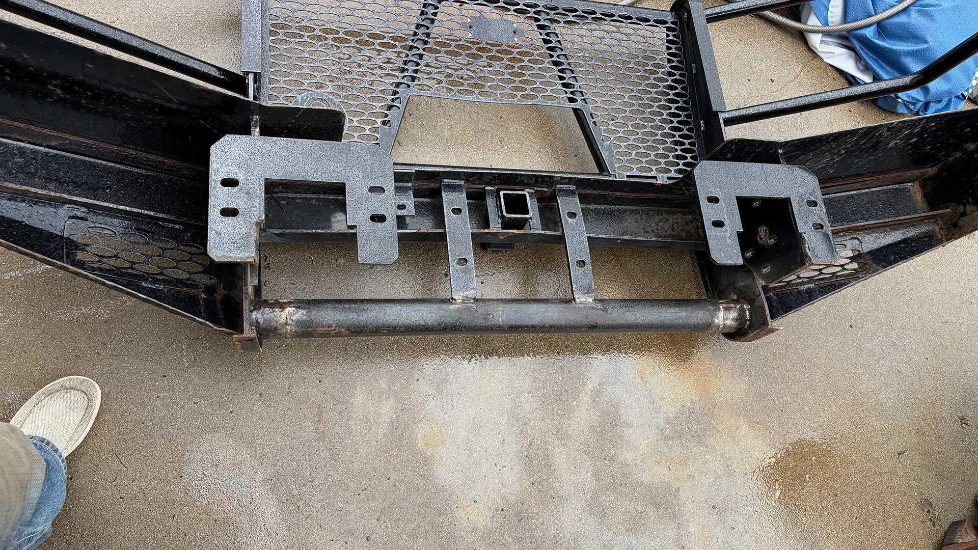 Metal frame structure with a ladder and grille, mounted on a concrete surface, with a person's left foot and a shoe visible at the bottom left corner, and a blue bag and black wheelbarrow in the background.