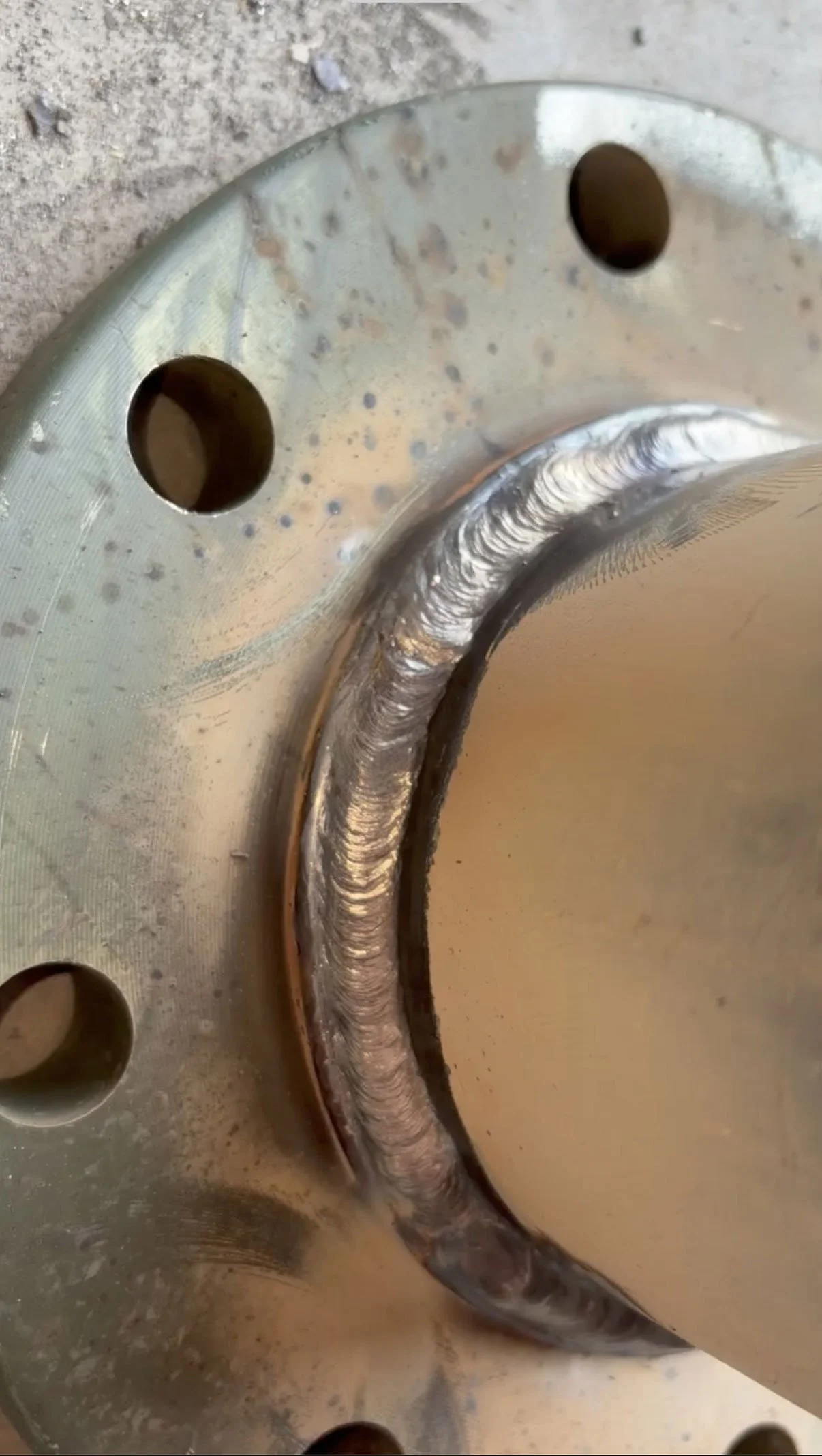 Close-up of a metallic machine part with a welded seam, multiple holes, and a textured, finished surface.