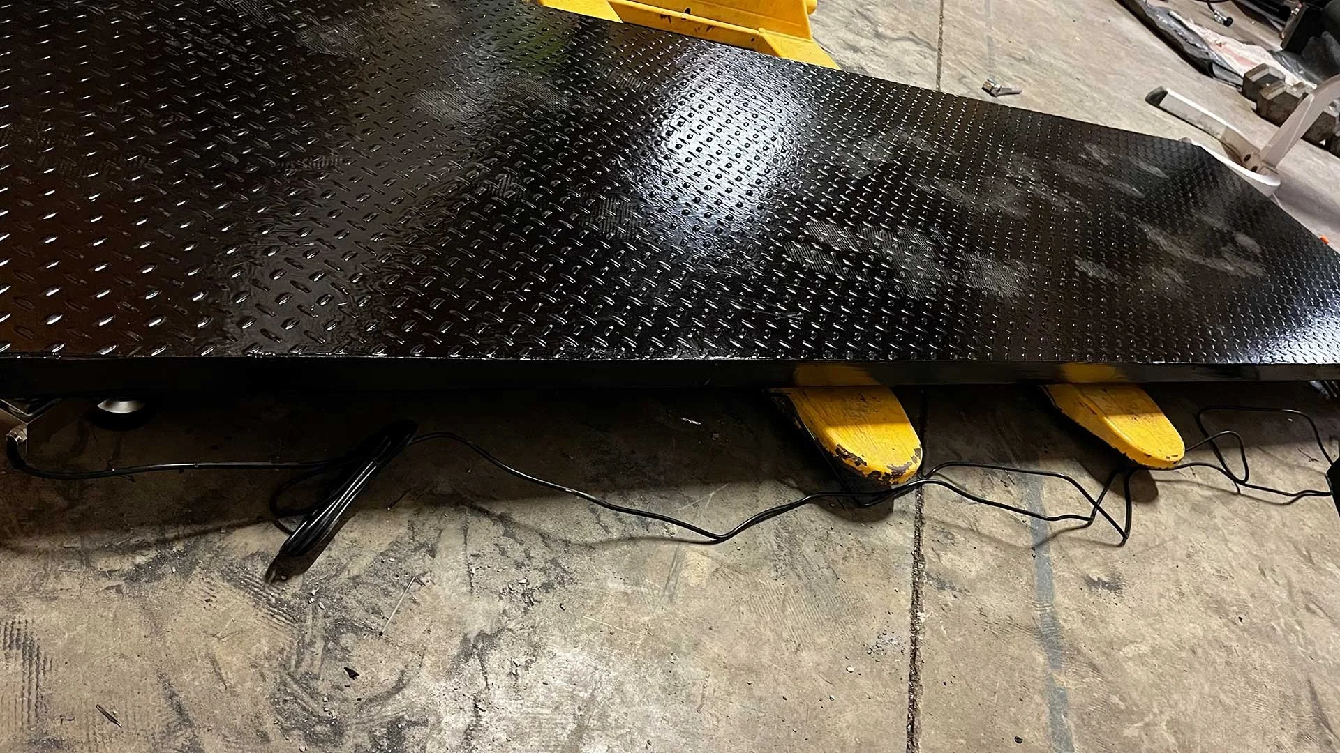 A black textured metal platform with a glossy finish, resting on yellow support beams, and electrical cords underneath on a concrete floor.