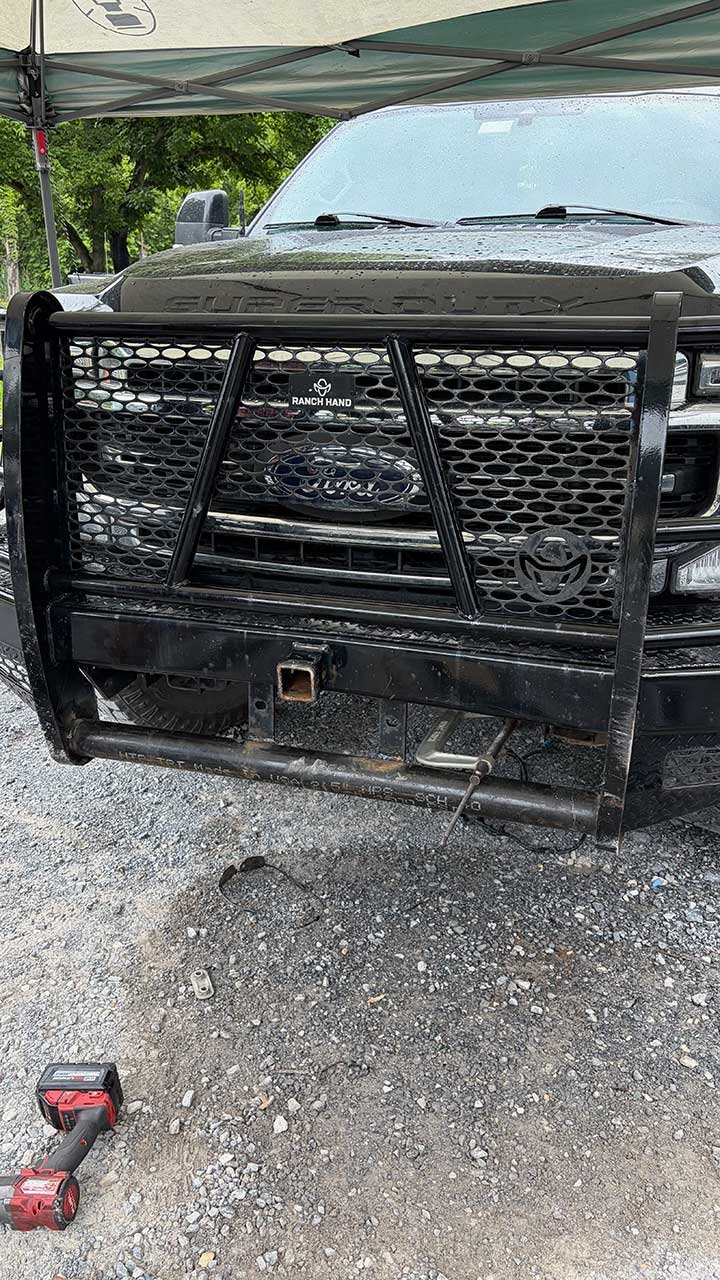 Front of a black pickup truck with a push bumper attached, parked on gravel, under a canopy. There is a small red and black power tool and a metal part on the ground in front of the truck.