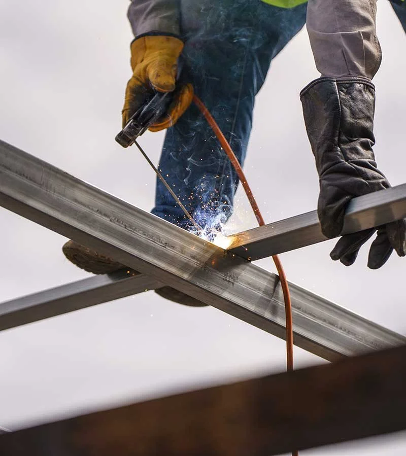 A person wearing protective gloves and boots welding metal beams together with sparks flying.