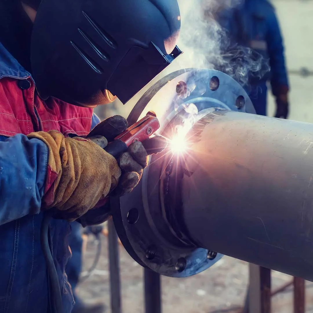 A person wearing a welding helmet and gloves is welding a large metal pipe with a visible bright spark and smoke.