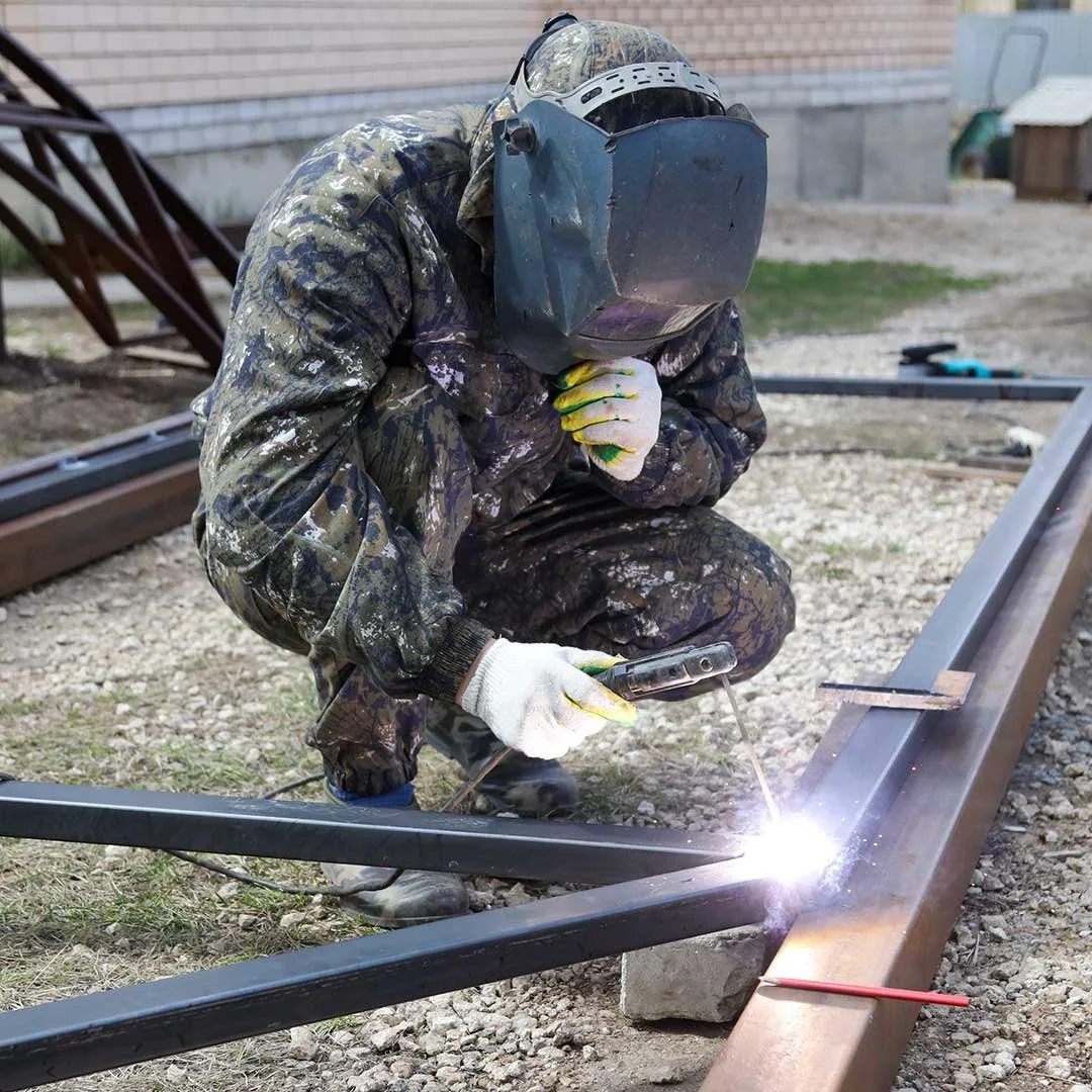 A person wearing protective gear, including a welding helmet and gloves, welding metal railway tracks outdoors on a gravel surface.