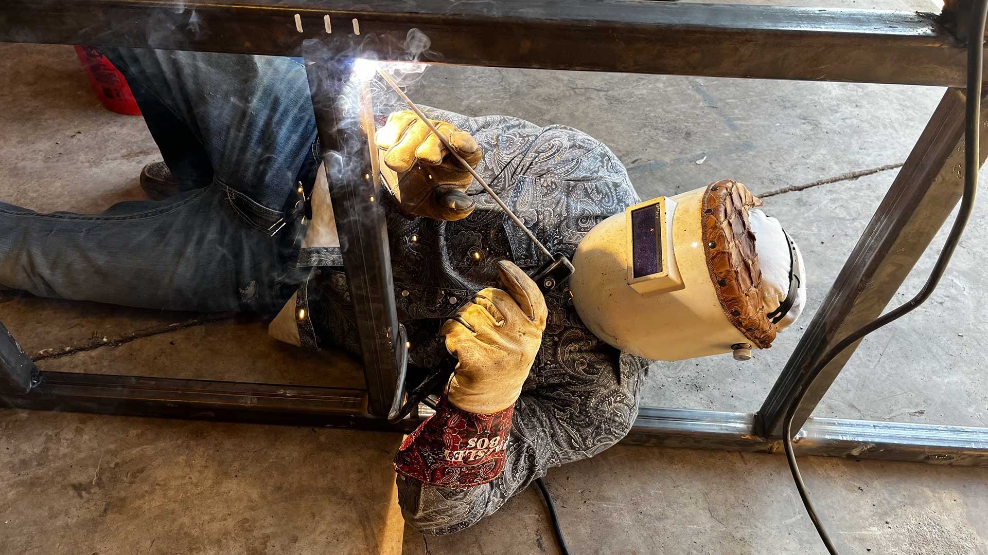 A welder lies on the ground welding steel under a worktable. They are wearing protective gloves and a welding helmet resting on their head, with welding sparks visible.
