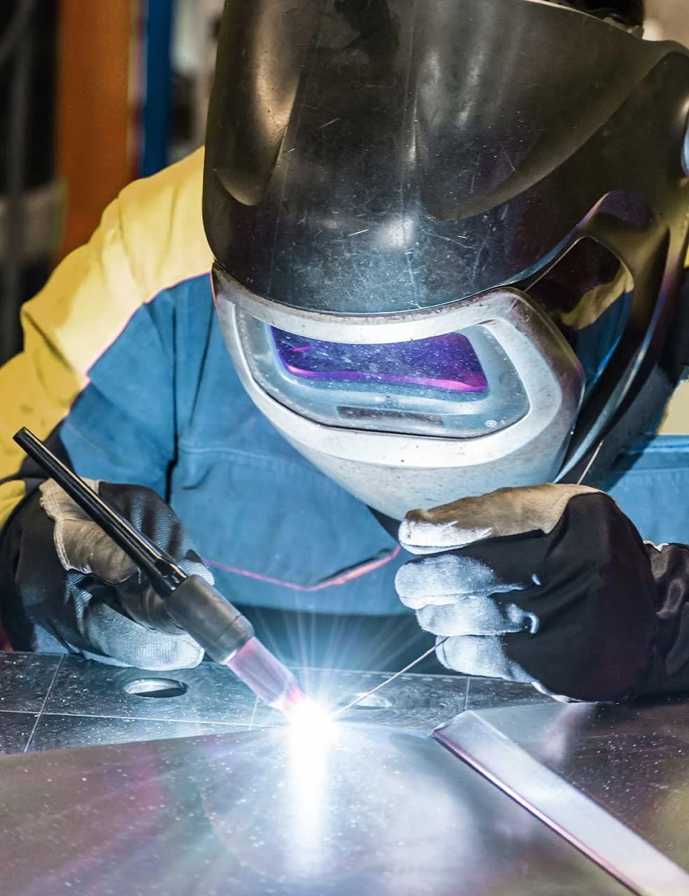 A person wearing a welding helmet and gloves is welding metal, with bright sparks and a glowing arc of light.