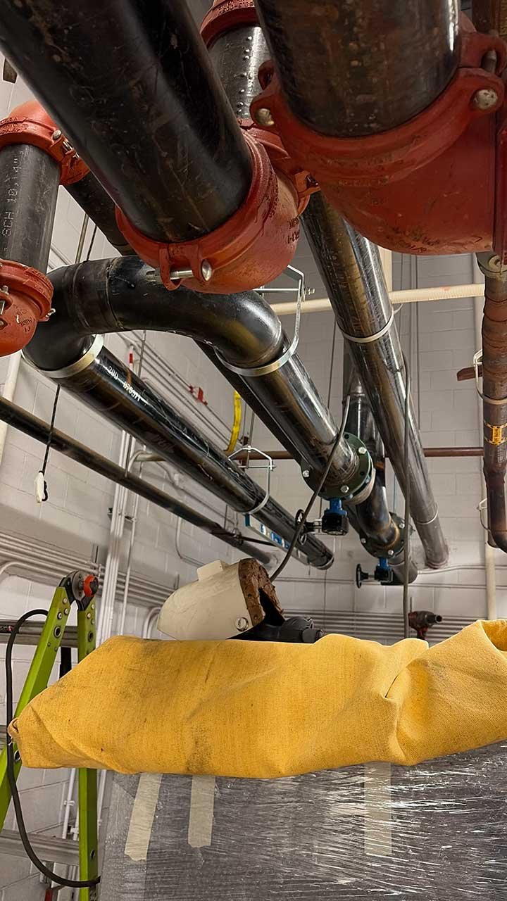 View of industrial pipes and valves overhead, with a yellow fabric cover or cloth in the foreground, and a ladder to the left.