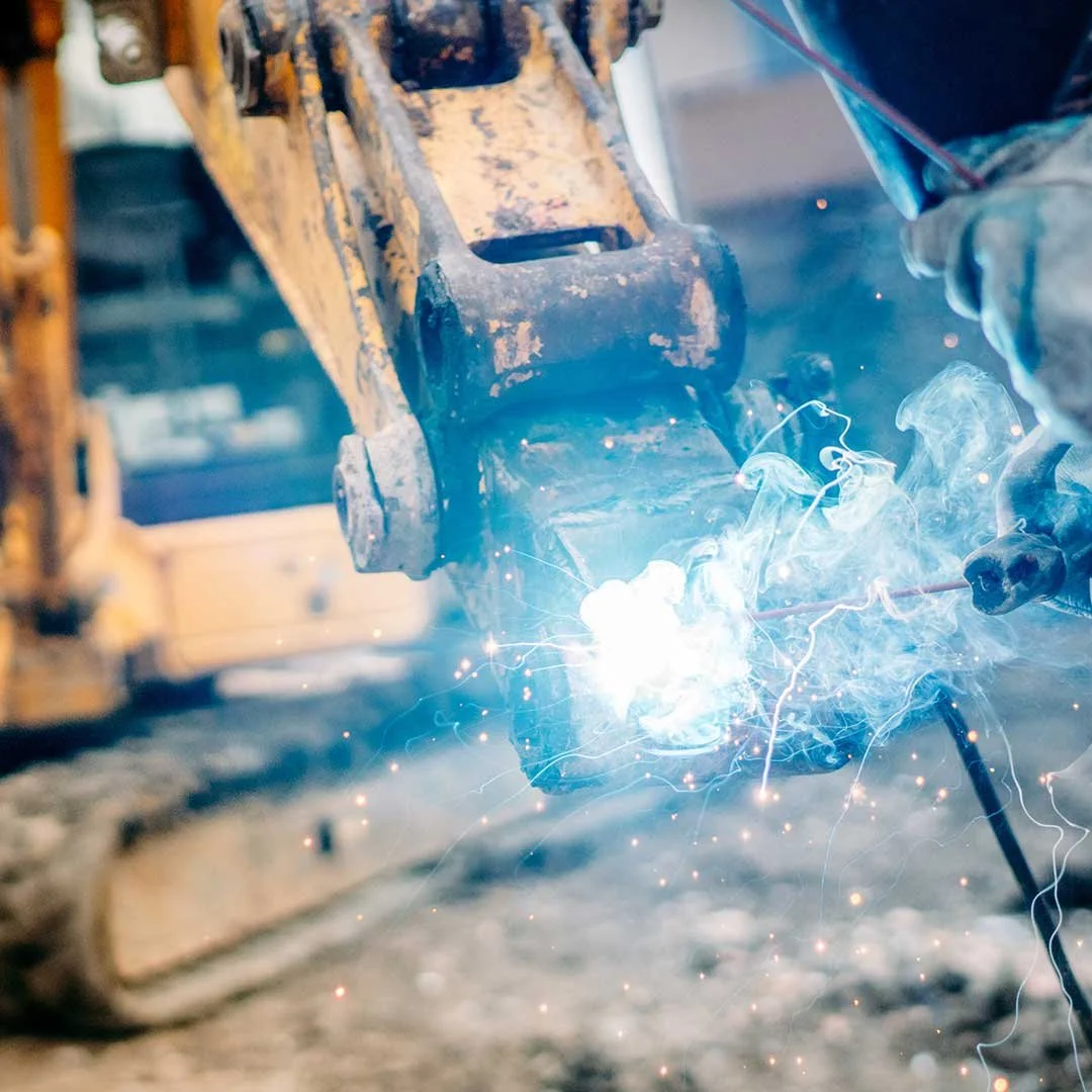 Close-up of a welding process on a yellow construction vehicle, with sparks and bright sparks visible, and a welder working on metal.