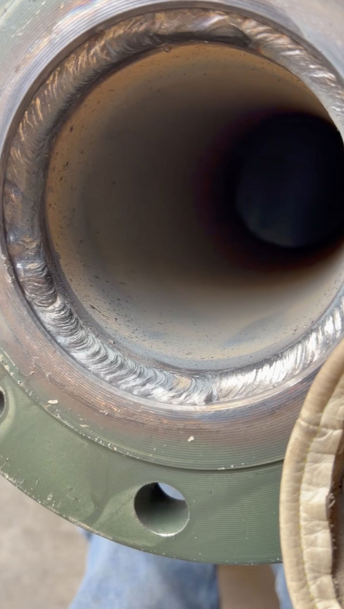 Close-up of a cylindrical metal component with visible welding beads around the edge, placed on a green surface with a hole.