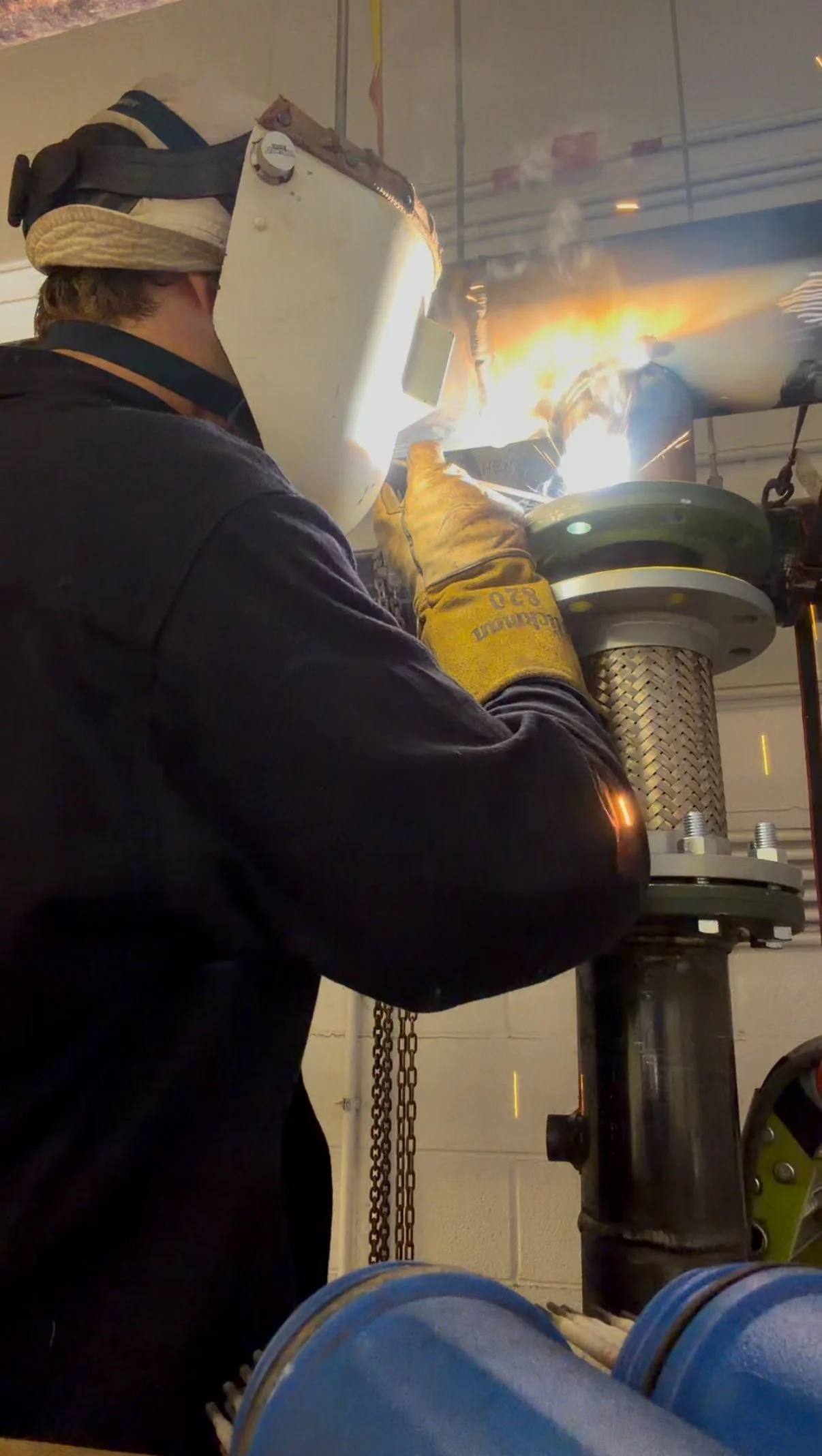 A person welding in a workshop, wearing a protective helmet and gloves, with bright sparks and a fiery glow from the welding process.