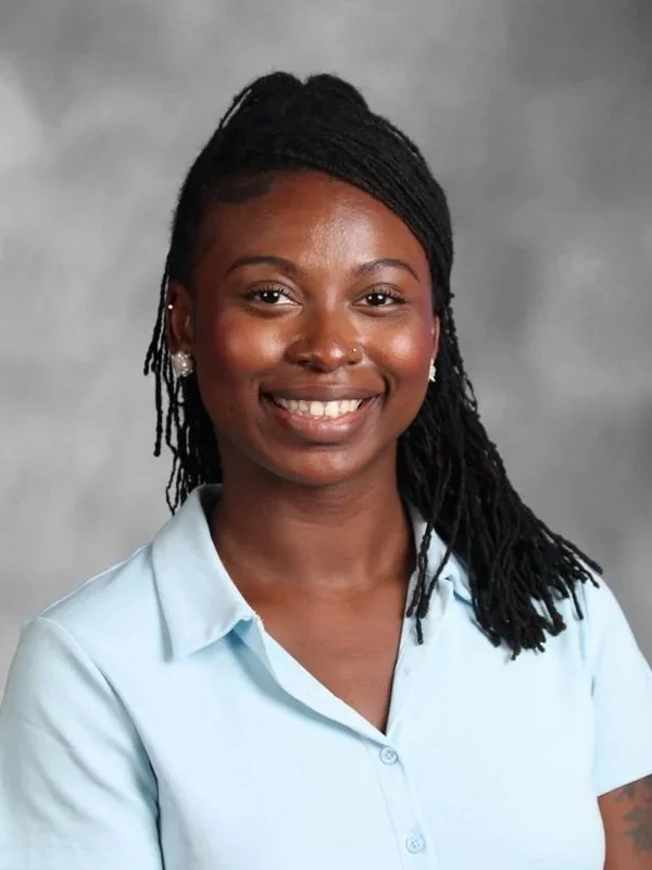A young African American woman with braided hair, wearing a light blue collared shirt and pearl earrings, smiling against a gray background.