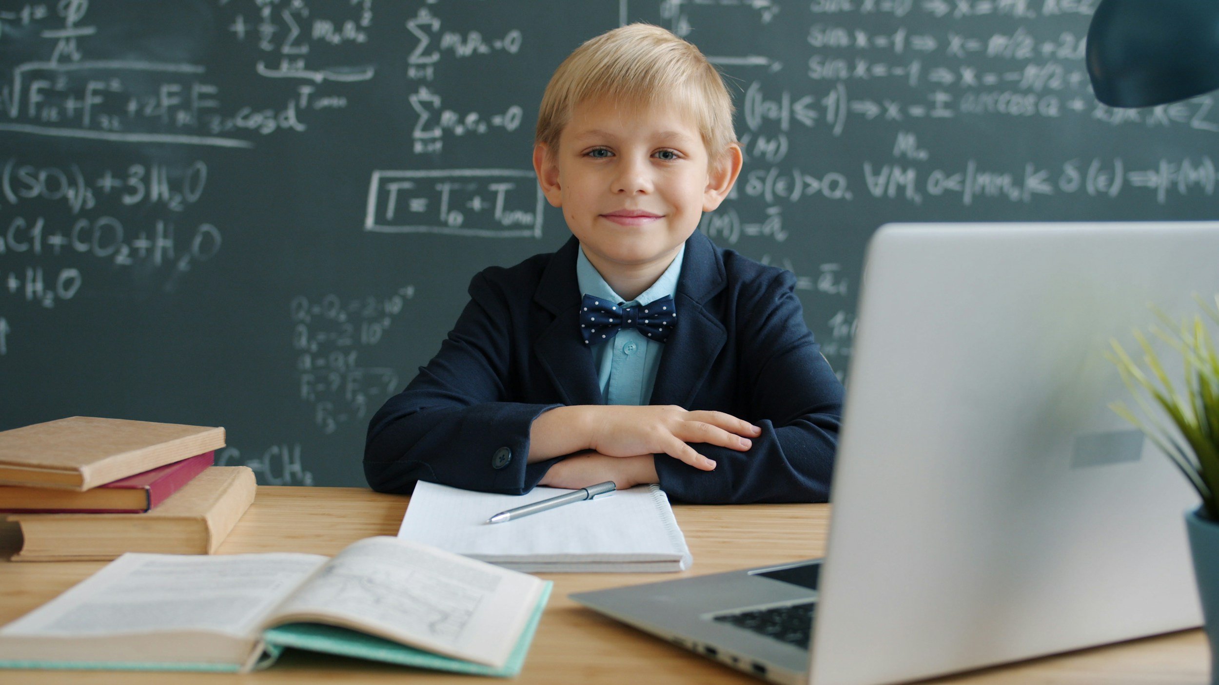 A young boy with blond hair, wearing a dark blazer and a polka dot bow tie, sitting at a desk in front of a chalkboard filled with mathematical equations, in a classroom setting, smiling at the camera.