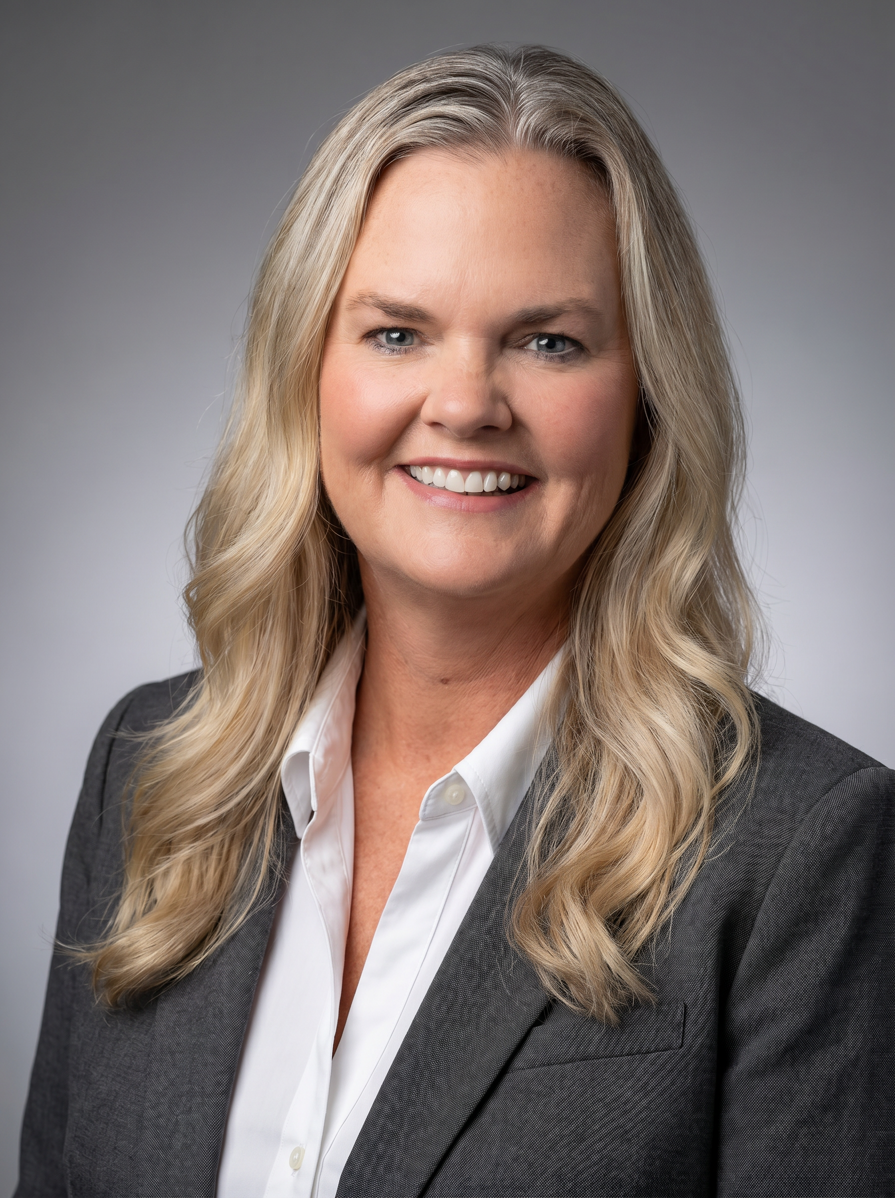 Professional woman with long blonde hair, wearing a gray blazer and white blouse, smiling against gray background.