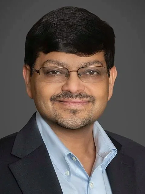 Portrait of a man with black hair, glasses, and a light blue shirt, smiling and wearing a dark blazer against a dark background.