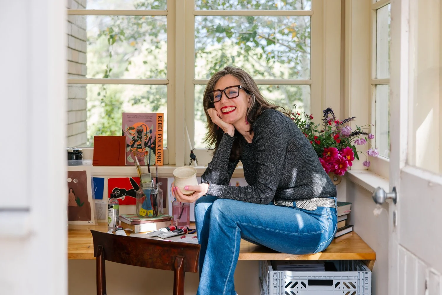 A woman with glasses and red lipstick sitting on a wooden desk, smiling, holding a cup, in a room with large windows and colorful flowers behind her.