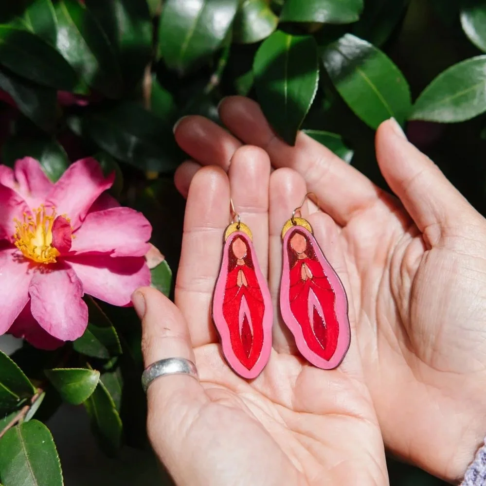 A person holding colorful, layered earrings resembling stylized woman figures with hands clasped in a prayer pose. The background includes green leaves and a pink flower with yellow stamens.