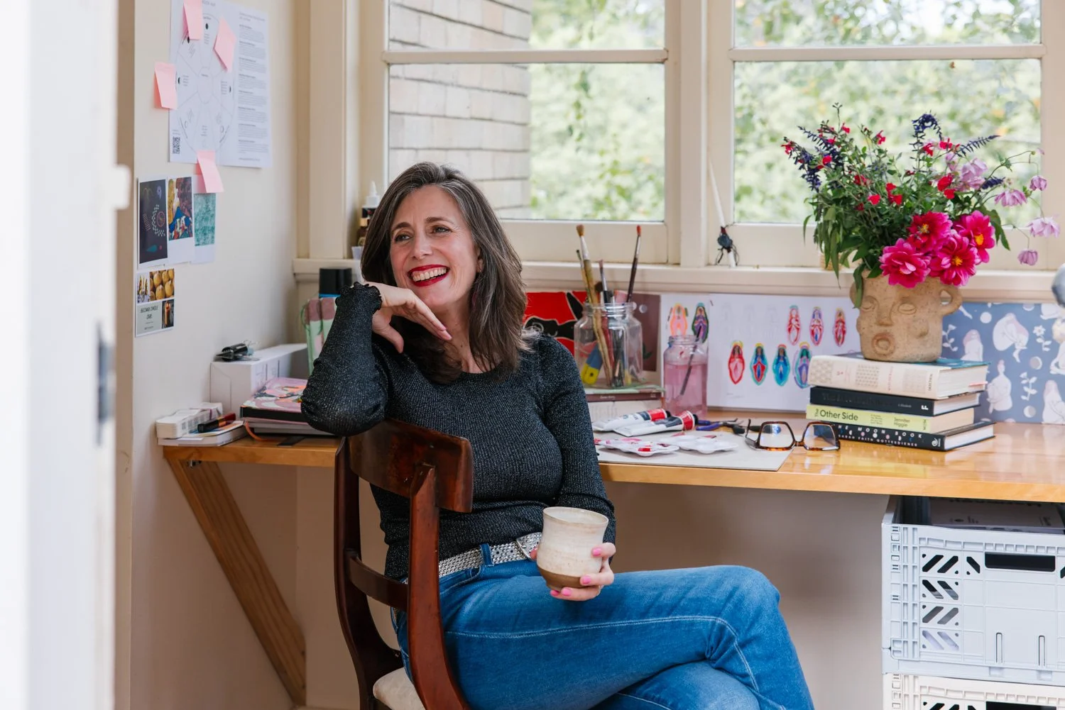 A woman with shoulder-length brown hair, wearing a black long-sleeve shirt and blue jeans, smiling while sitting on a wooden chair in a cozy, well-lit home office or craft room. She holds a ceramic cup in her hand. The room has a desk with art supplies, books, a large vase with colorful flowers, and windows with a view of green trees outside.