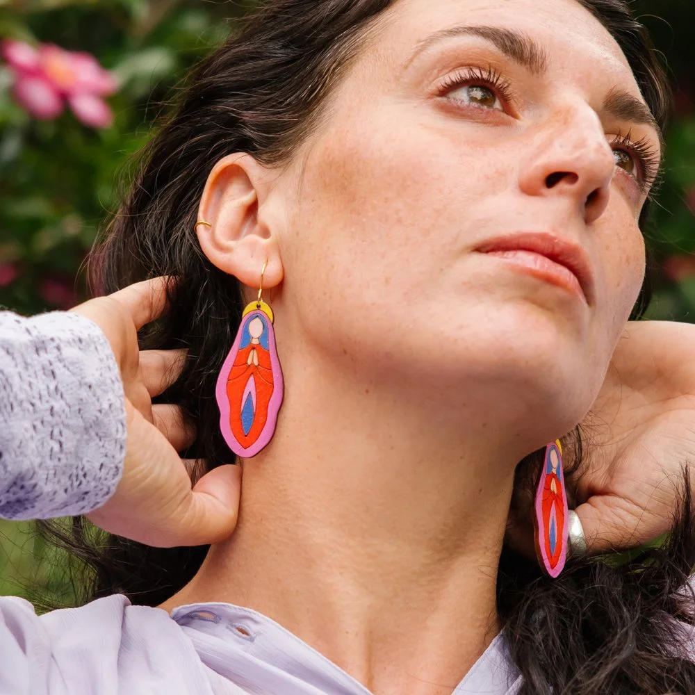 A woman with dark hair and freckles wearing colorful abstract earrings, outdoors with pink flowers in the background.