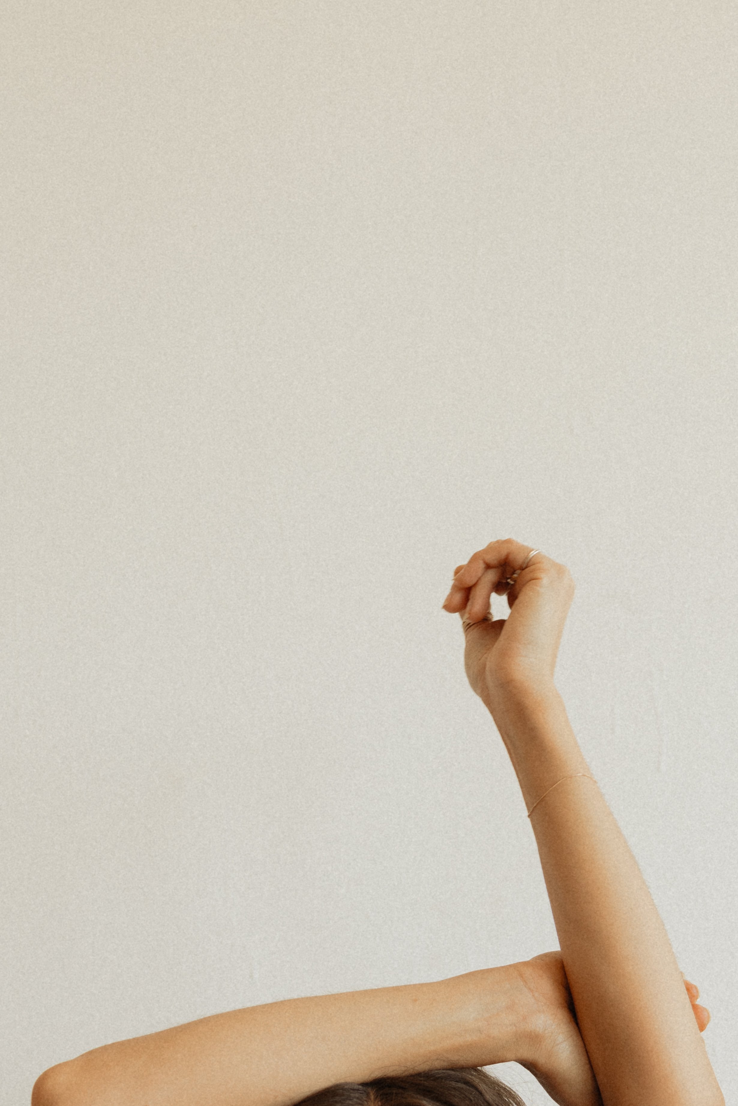 A person with long hair is stretching their arms above their head against a plain beige wall.