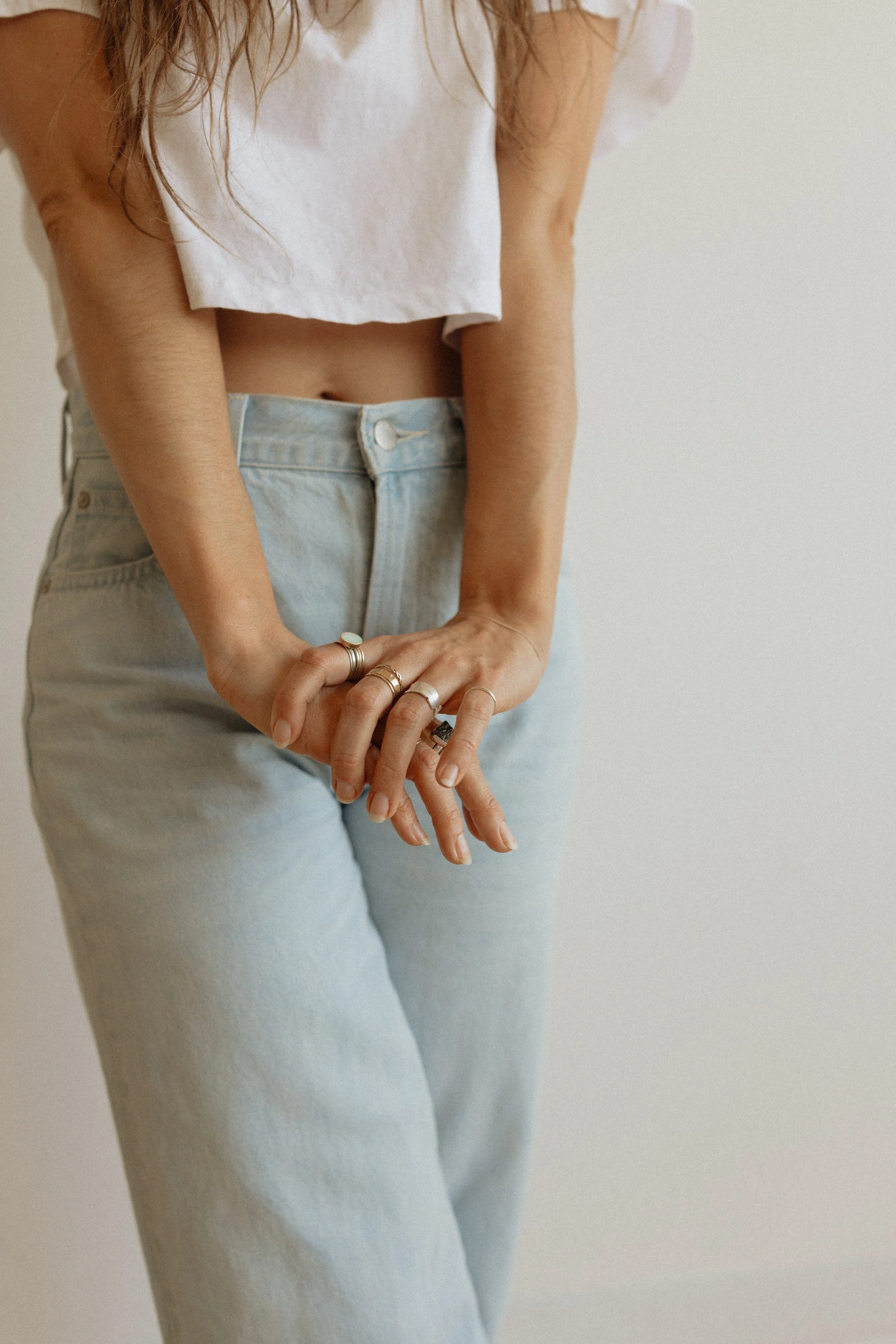 Close-up of a woman wearing a white cropped top and light blue jeans, with multiple rings on her fingers, against a plain neutral background.