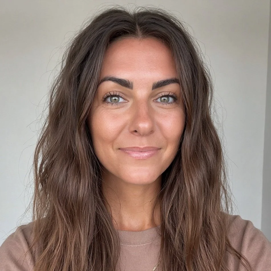 A woman with long wavy brown hair, fair skin, and light-colored eyes, smiling at the camera against a plain light background.