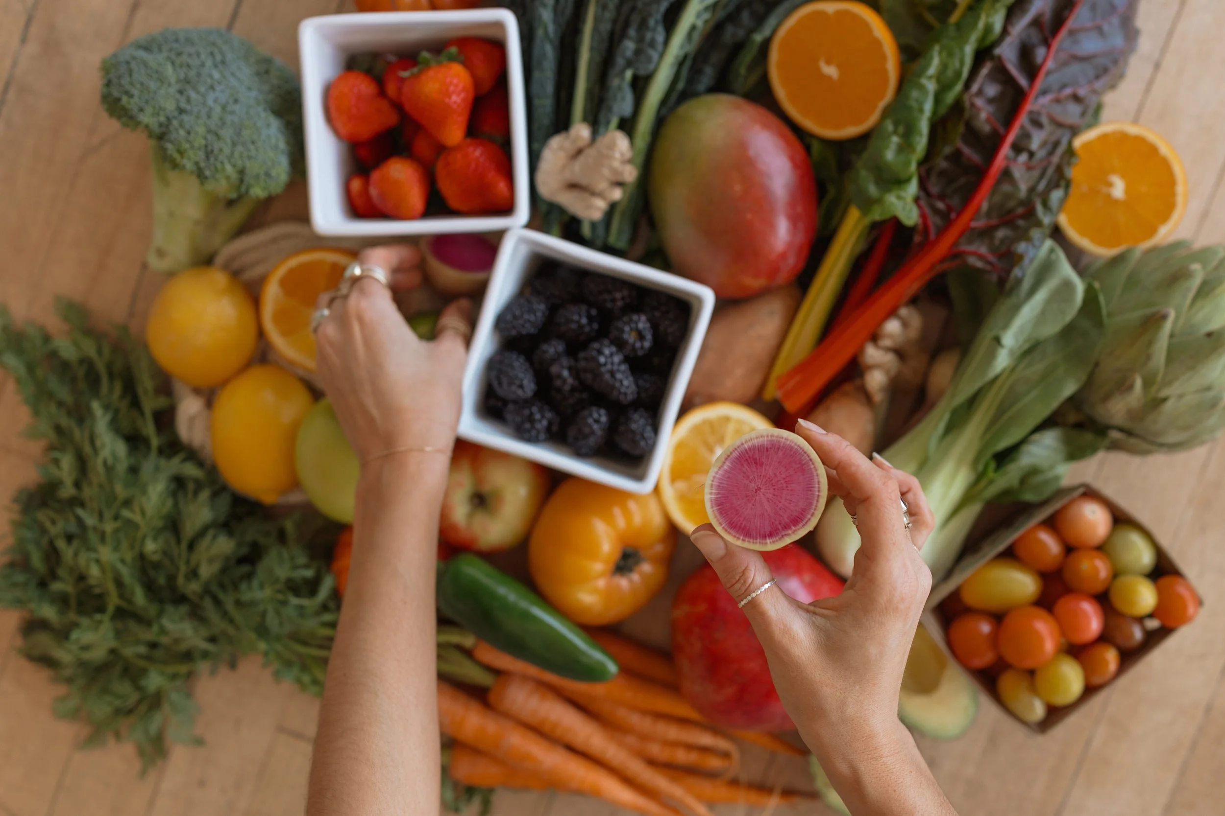 Hands holding slices of dragon fruit over an assortment of fresh fruits and vegetables, including strawberries, blackberries, oranges, tomatoes, carrots, cucumbers, apples, broccoli, and leafy greens.