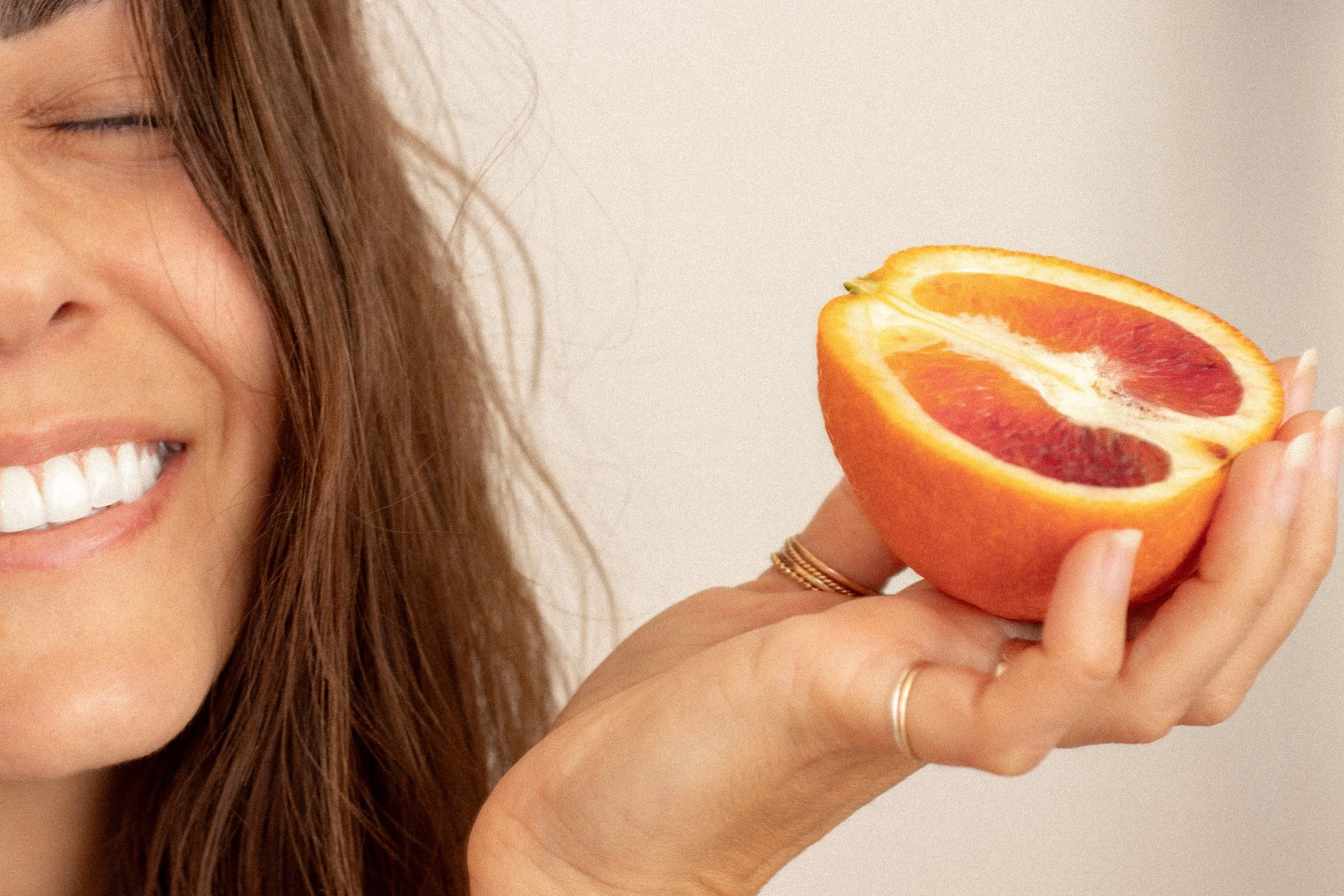 A woman with brown hair smiling and holding a halved grapefruit close to her face.
