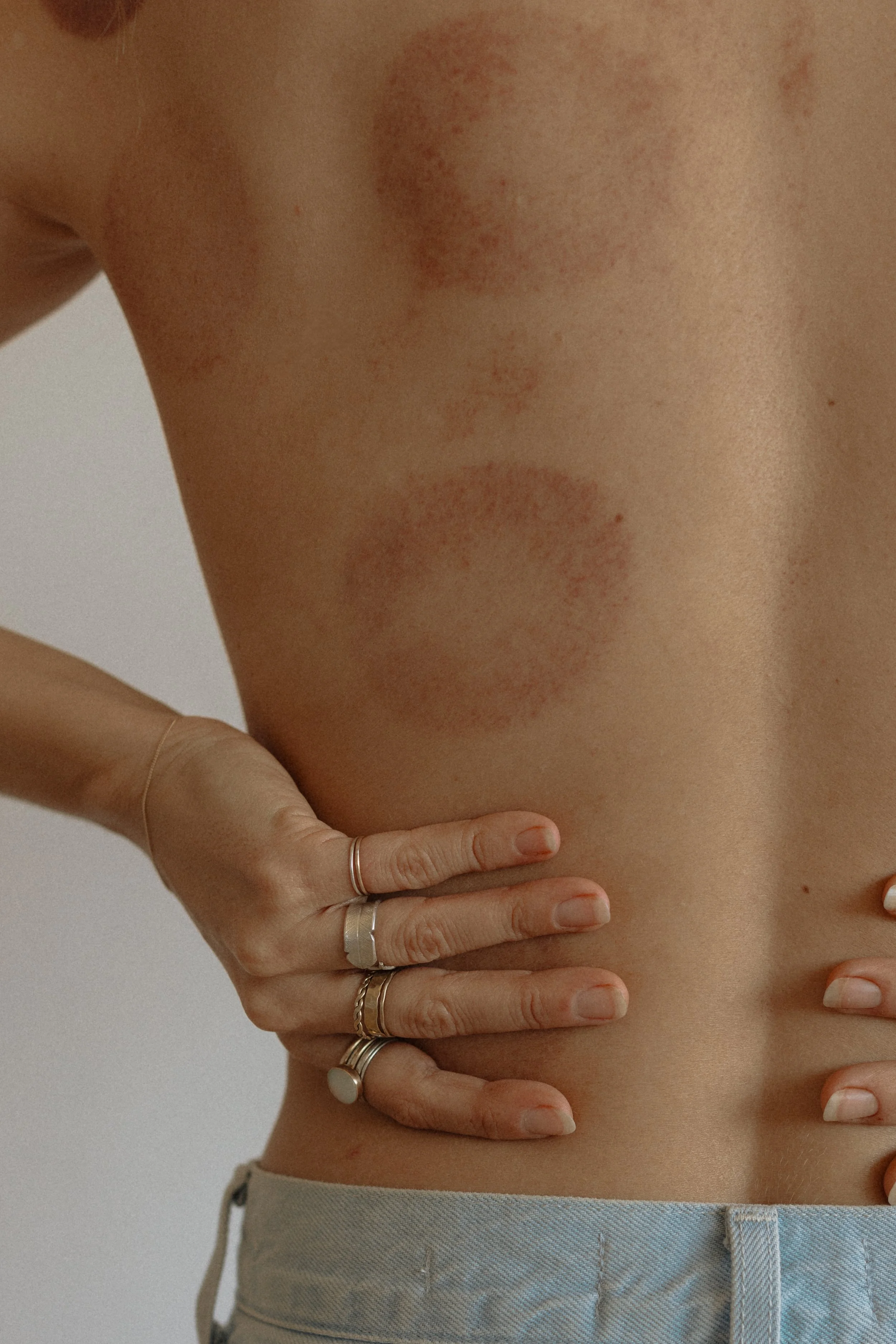 Close-up of a person's bare midsection with their hand resting on their stomach, showing multiple rings on fingers and a beige waistband of pants.