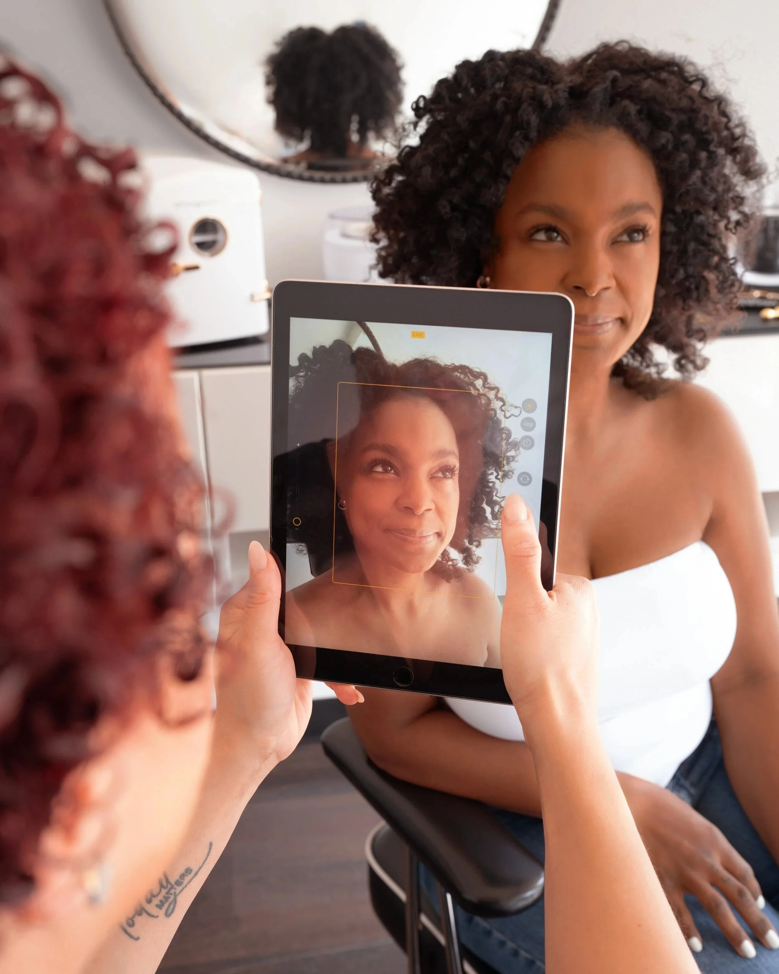 A woman with curly hair is taking a selfie of herself using a tablet. There is a mirror behind her reflecting her back, and she is sitting in a room with white walls and a white device in the background.