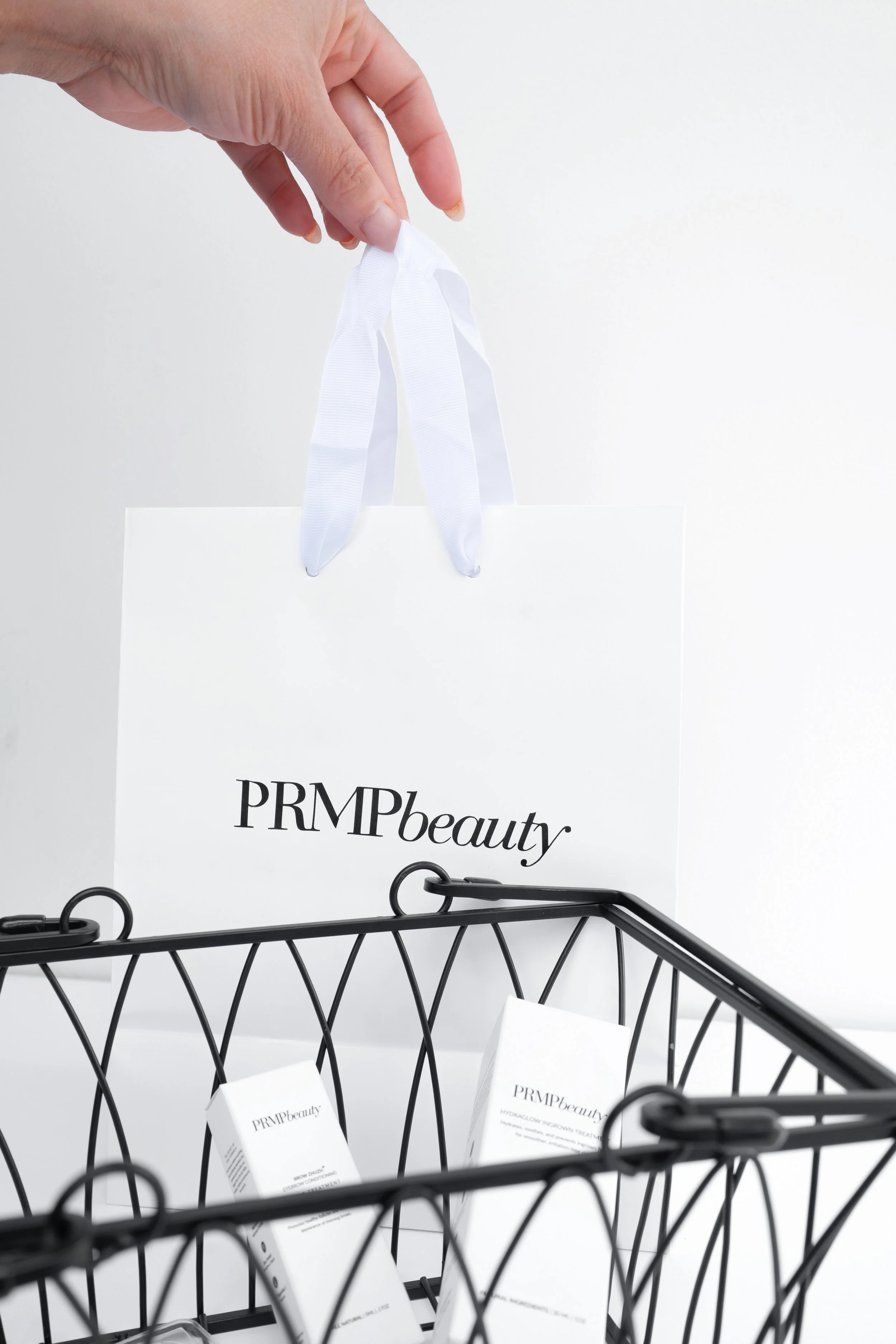 A hand is holding white ribbon attached to a shopping bag with PRIMPbeauty printed on it. There are similarly branded skincare products in a wire basket below.