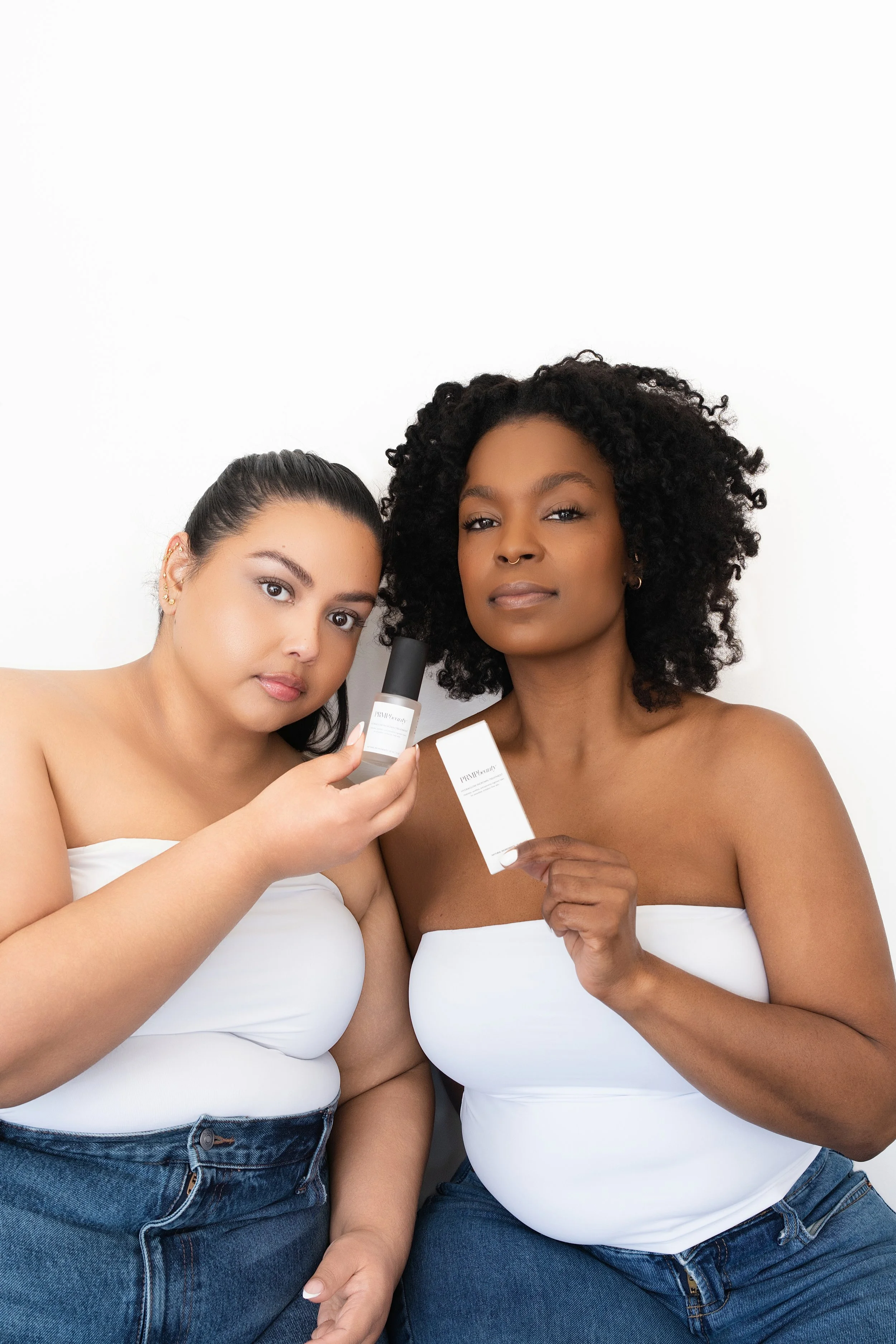Two women, wearing white tank tops and jeans, sit close together against a plain white background. The woman on the left has light skin, dark hair pulled back, and multiple earrings, and she is holding a bottle and a box of skincare products. The woman on the right has darker skin, curly black hair, and a septum piercing, and she is holding a box of the same skincare product.