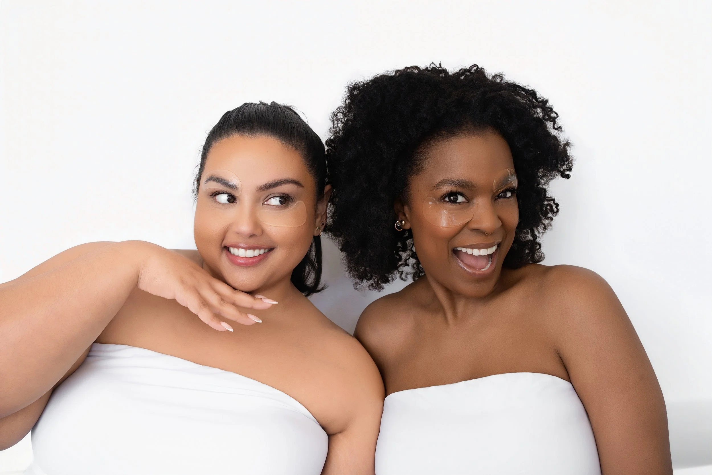 Two women with toned bodies, wearing white tube tops, pose for a photo against a plain white background. The woman on the left has dark hair in a high ponytail and is smiling, while the woman on the right has curly hair and is making a surprised or joyful expression. Both have under-eye patches on their faces and are shirtless below the tops.
