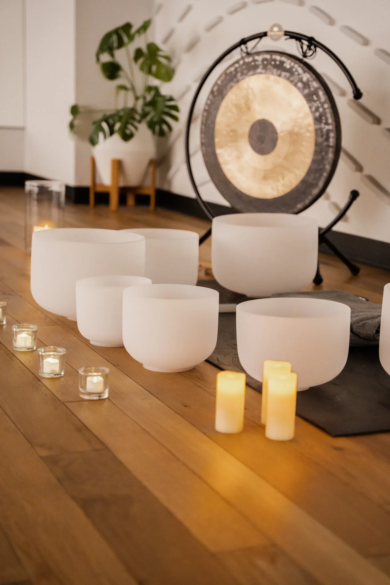 A gong with a black frame and a golden surface is positioned behind cream-colored bowls on a wooden table, with glowing candles around.