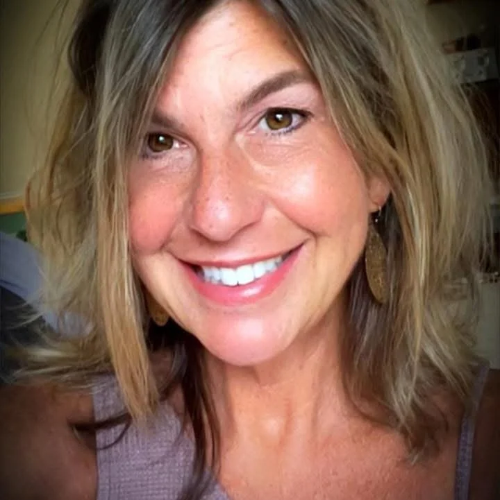 Close-up of a smiling woman with wavy blonde hair and brown eyes, wearing gold earrings and a sleeveless top.