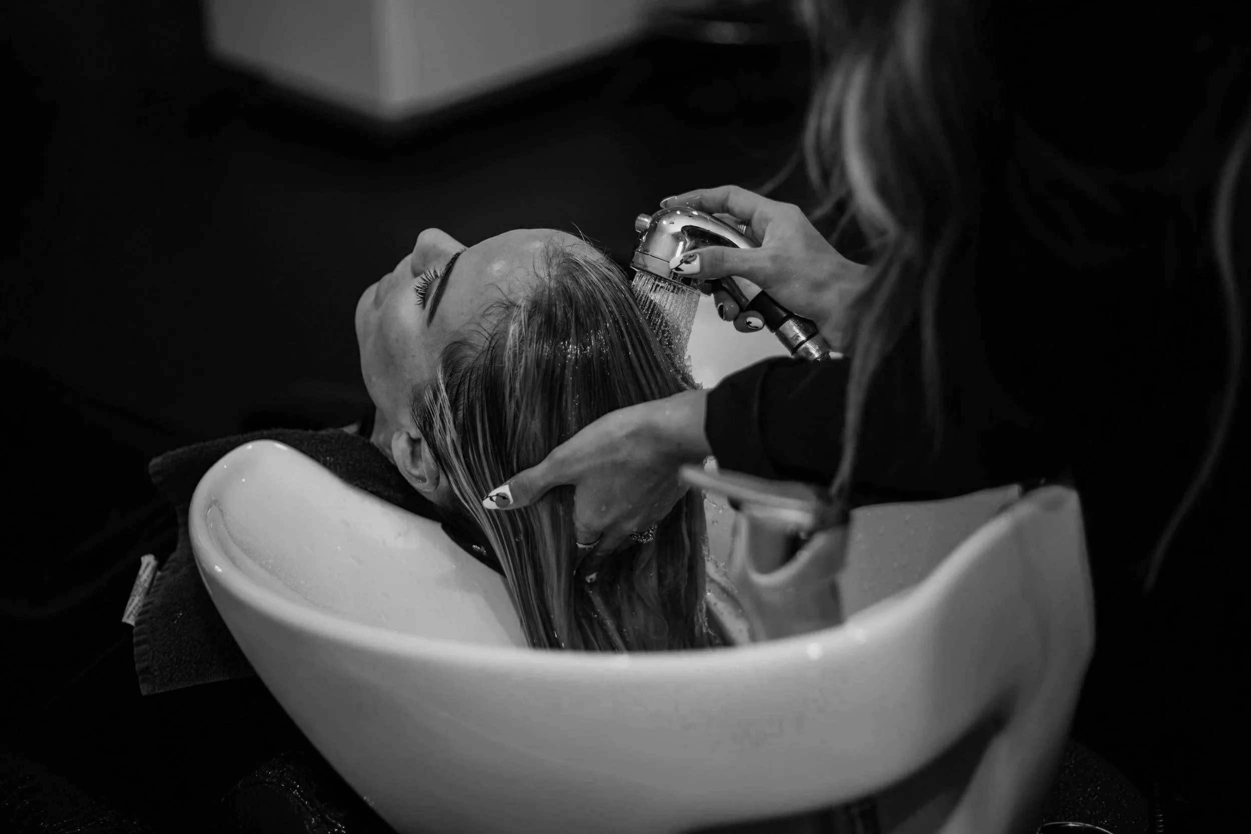 Black and white image of a woman with her head tilted back in a salon sink, receiving a hair wash, with water flowing from a handheld nozzle.