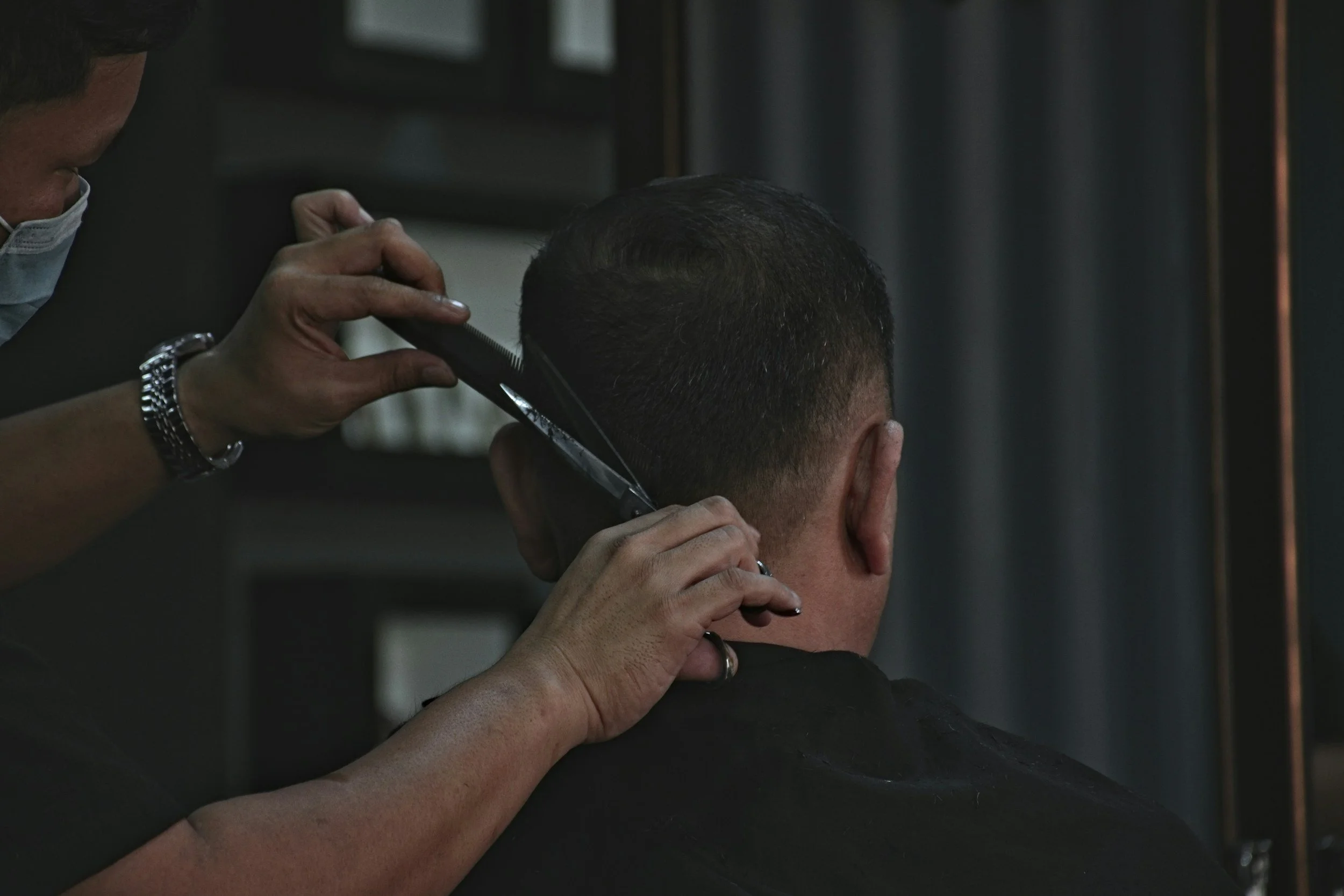 A barber trims a man's hair with scissors and comb, wearing a face mask in a barbershop.
