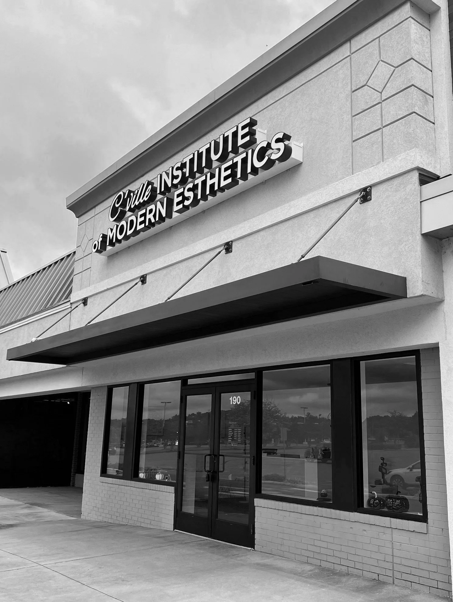 Front of a beauty or skincare institute with a sign reading 'Crille Institute of Modern Esthetics' in black and white. The building has large windows and a brick exterior, with the street and parking lot visible in the reflection.
