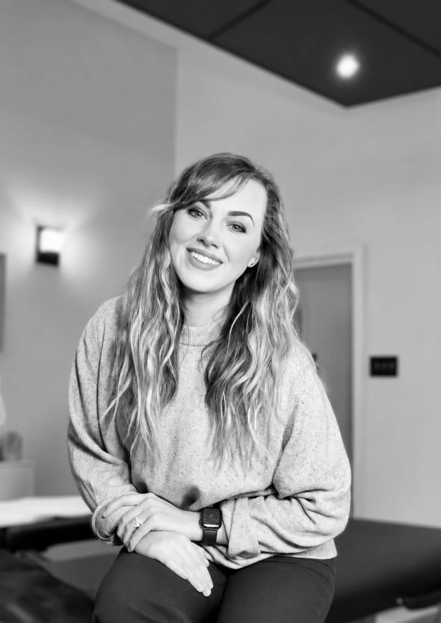A woman with wavy hair smiling at the camera in a room with modern lighting and minimal decor, in black and white.