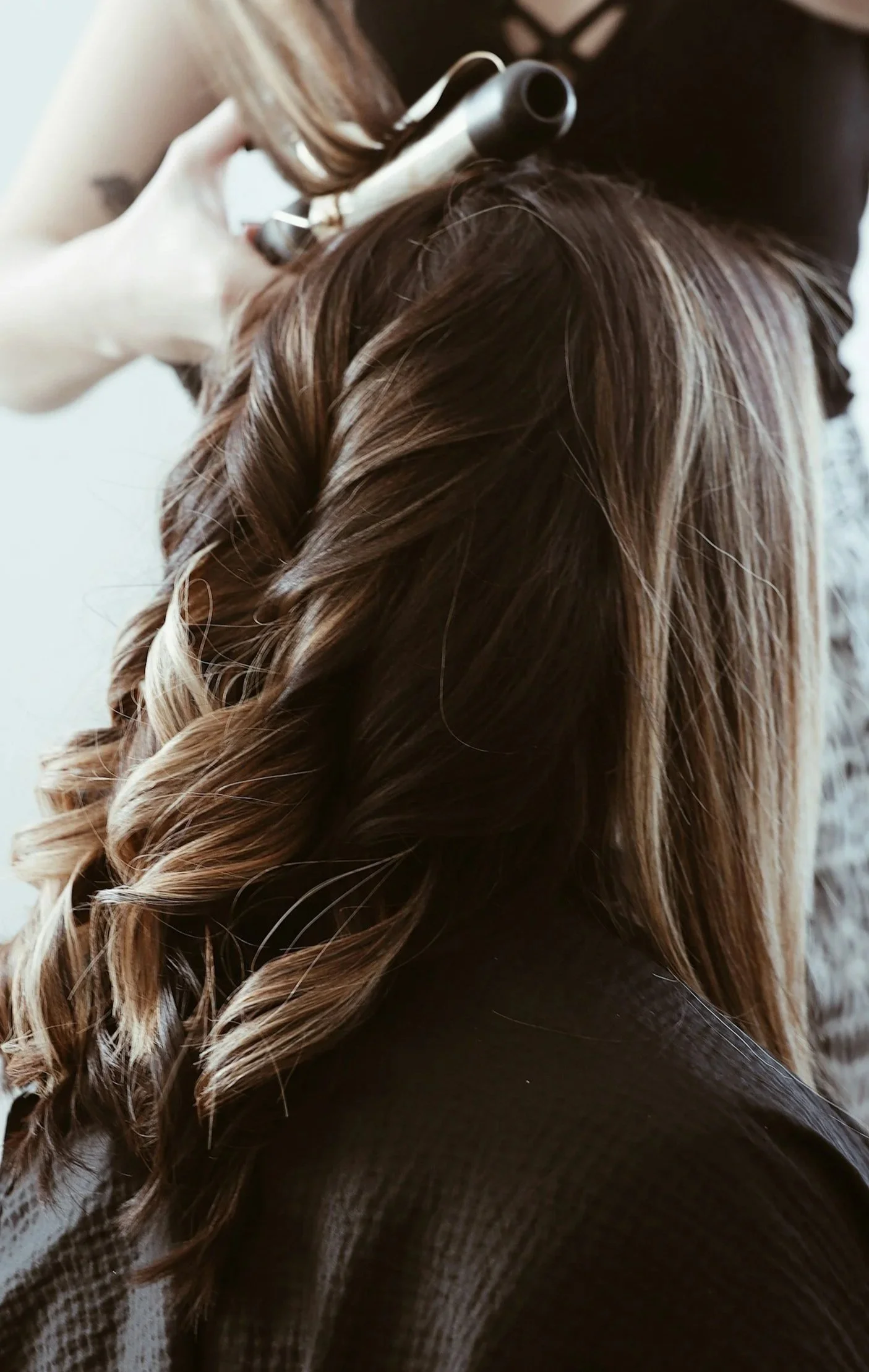 Close-up of a woman with long, wavy brown hair wearing a black top and a checkered coat, holding a curling iron to her hair.