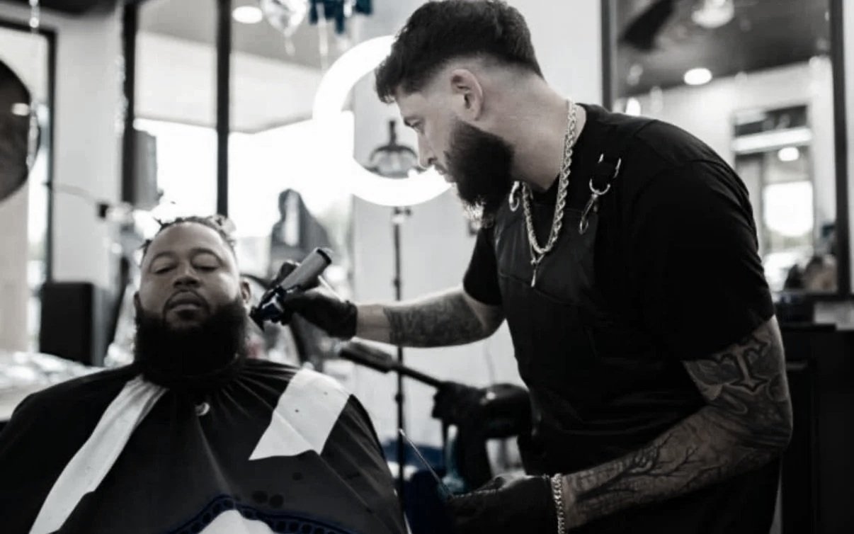 A man getting his beard trimmed at a barbershop by a barber with tattoos, wearing black gloves, a chain necklace, and dark clothing.