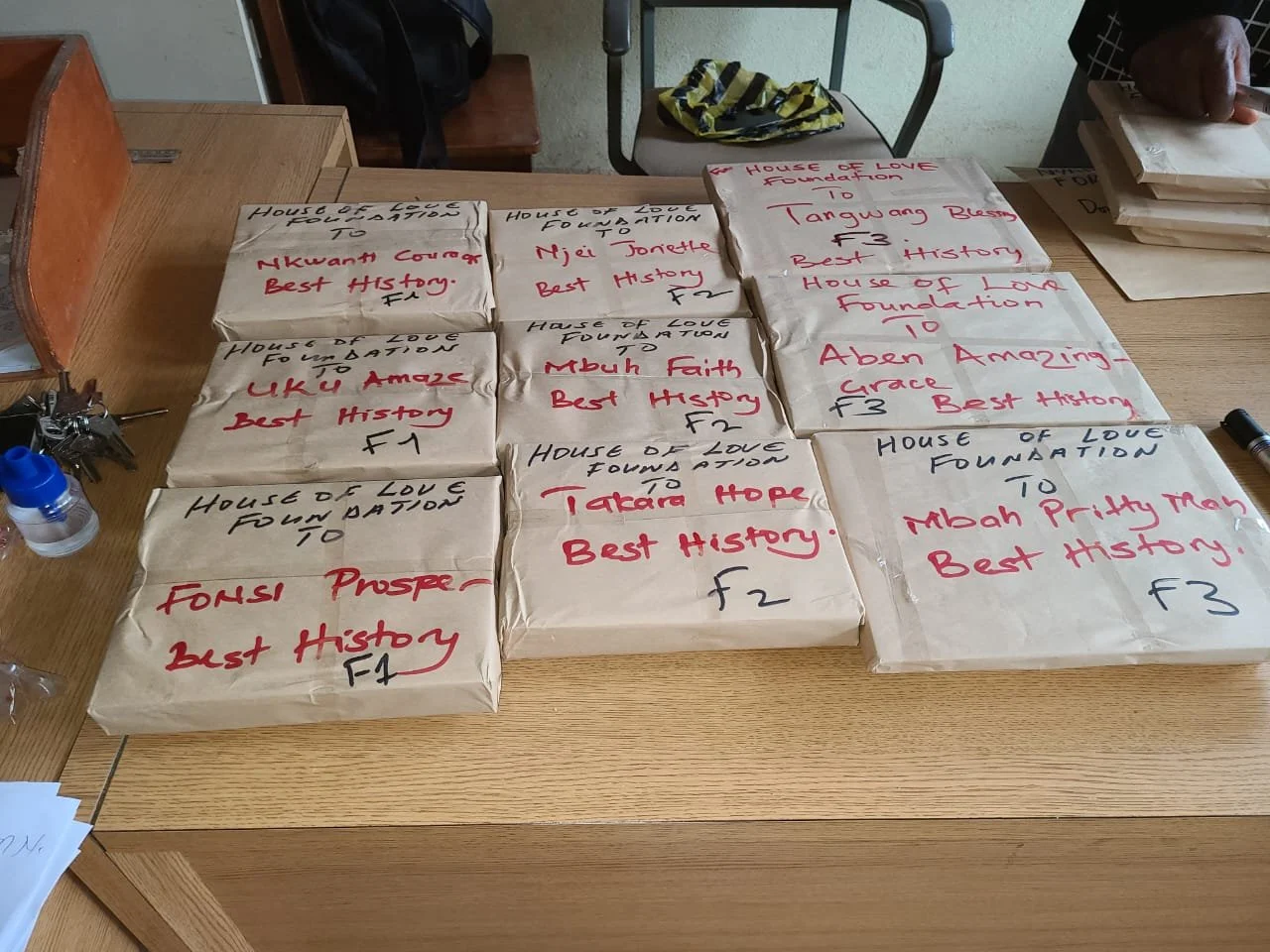 Multiple paper packages with handwritten labels on a wooden table, referencing the House of Love Foundation and honoring various individuals, with some packages marked 'Best History' and others with different dedications.