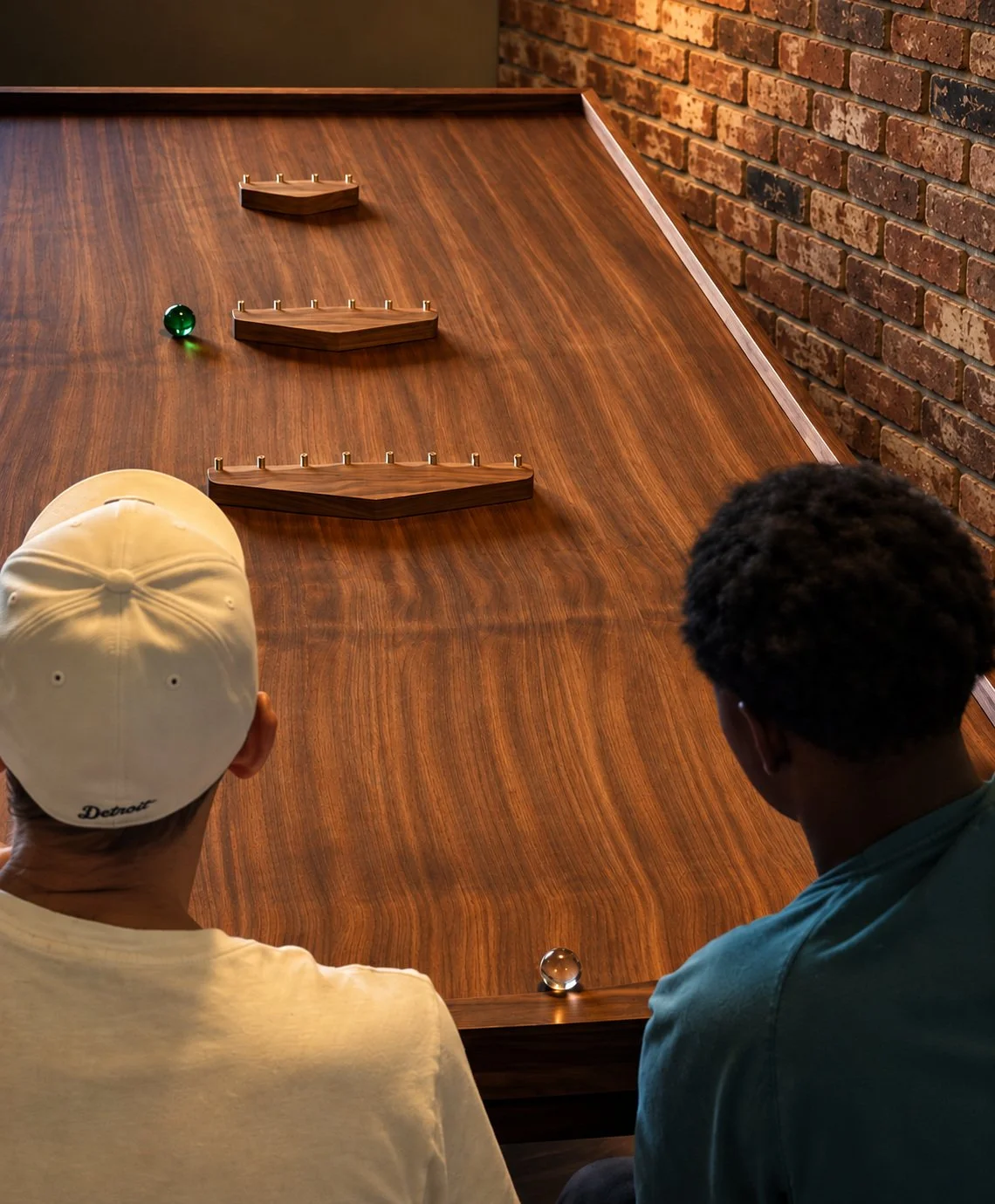 Two players look down a Vortex468 walnut game table beside a brick wall.