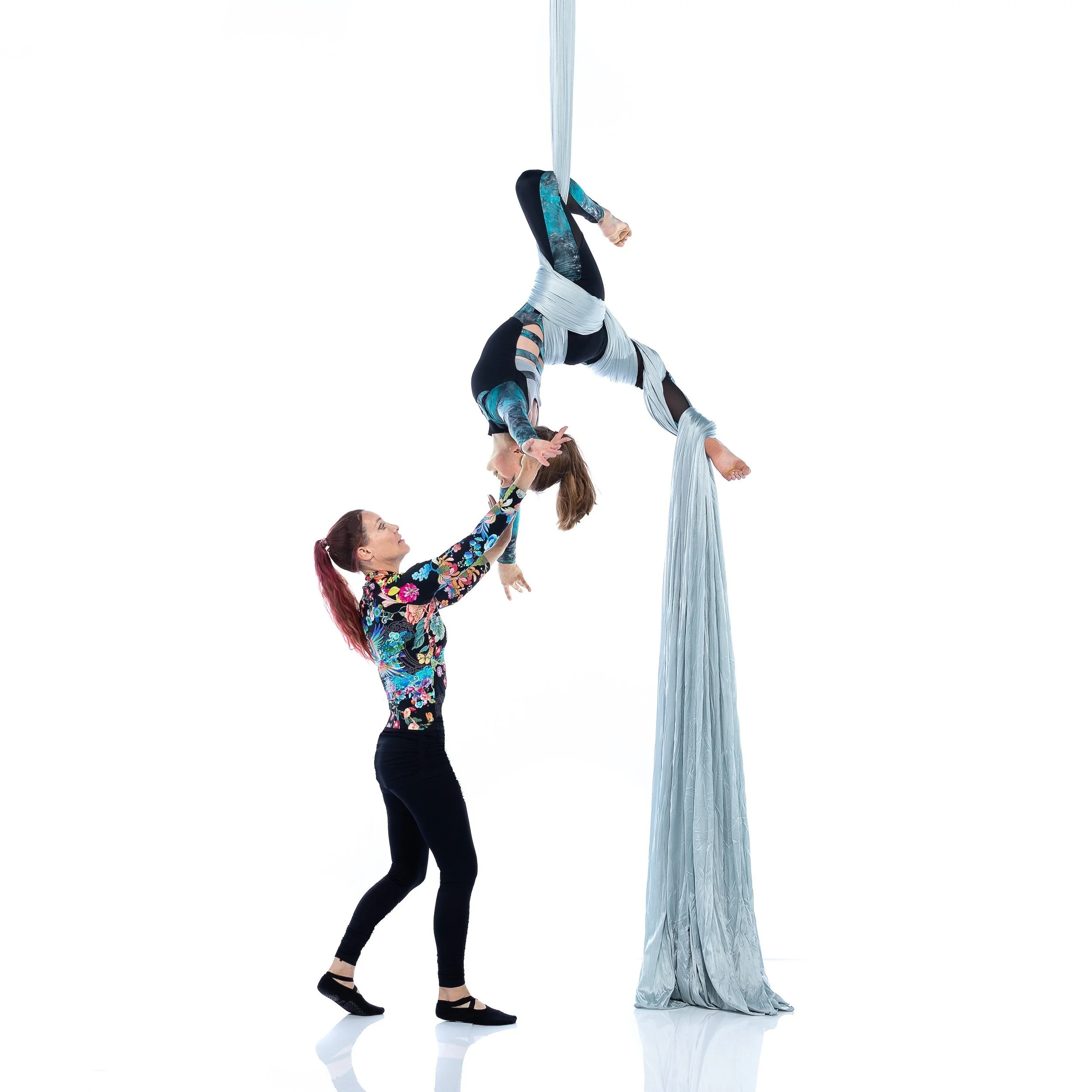Aerial silk performers demonstrate an acrobatic pose, with teacher supporting student who is upside down, both holding onto light blue silk fabric, against a plain white background.