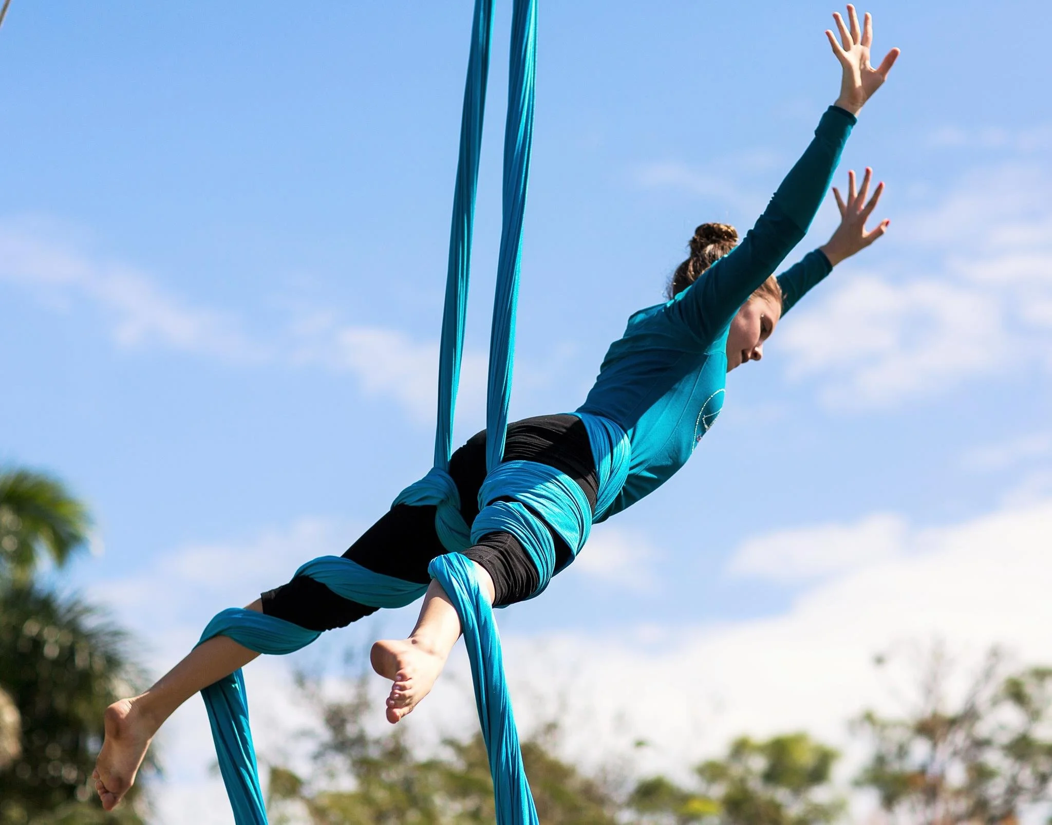 A woman in blue athletic clothing practicing aerial silk acrobatics outdoors against a blue sky, with trees partially visible in the background.