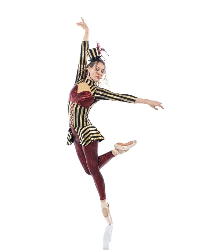 A ballet dancer in a colorful costume and makeup performing a pose with one arm raised and one leg lifted.
