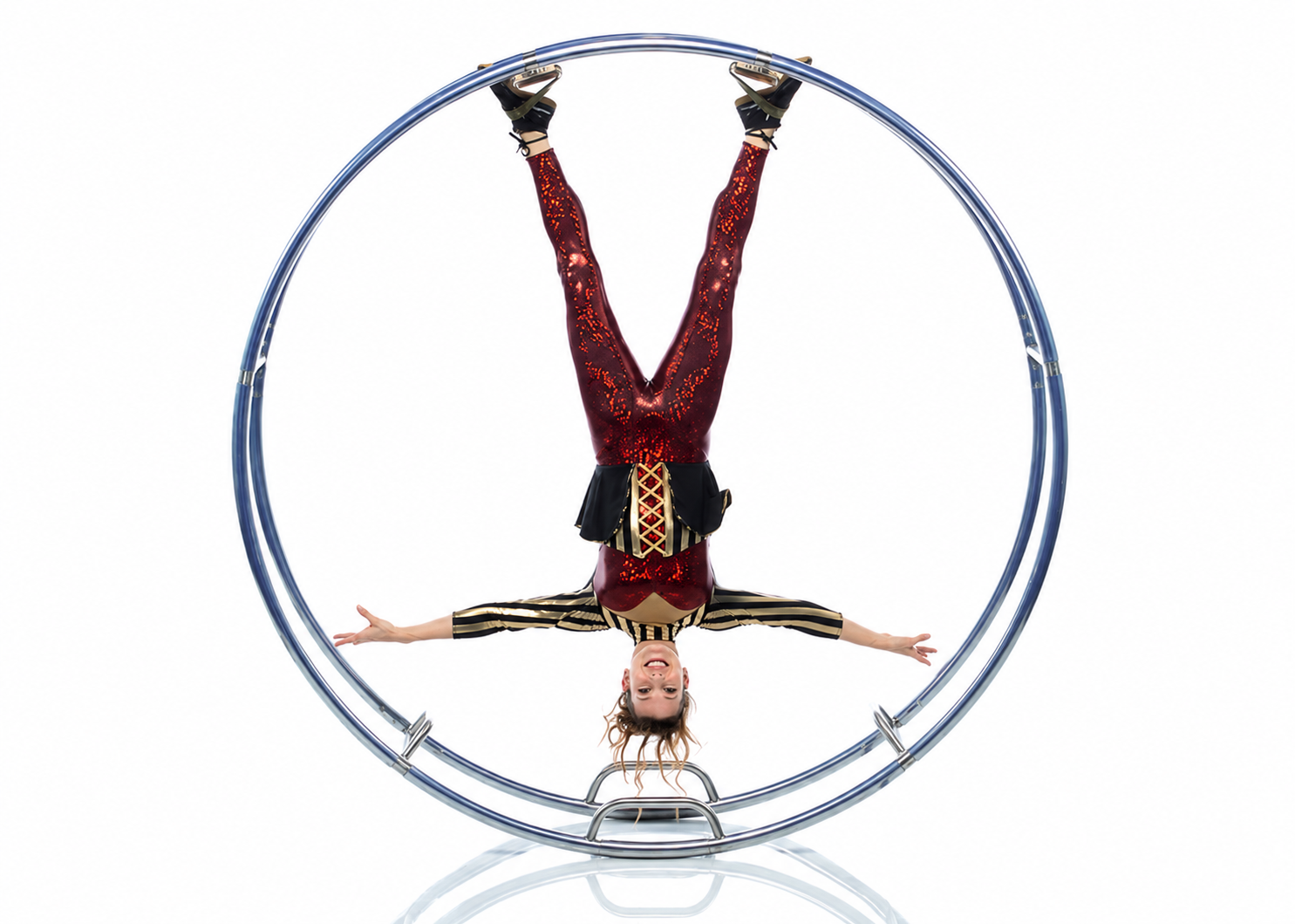 A young female acrobat is performing a handstand inside a German Wheel smiling at the camera, with arms extended to the sides and legs extended upward in a split.