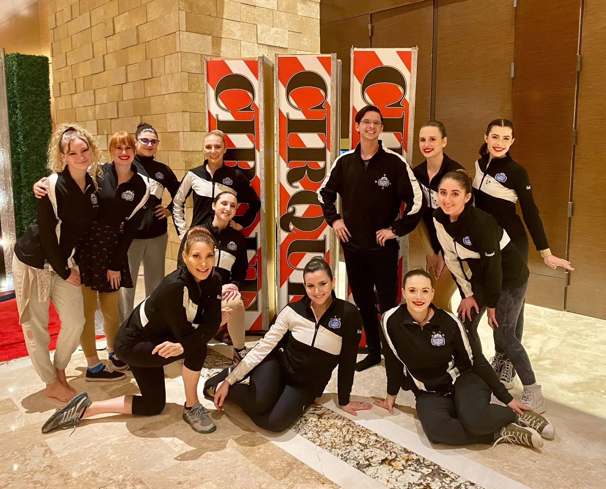 Group of ten young people, mostly women, posing together indoors in front of a large checkered sign that reads 'Circus'. They are all wearing black and white jackets with a logo, and some are squatting or kneeling while others are standing. The background includes a brick wall and wooden door.