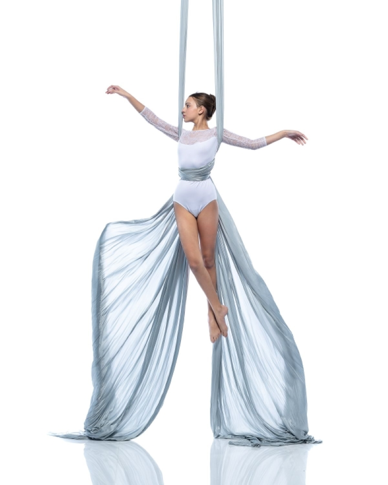 A woman performing an aerial silk routine, suspended from two long silver fabrics, with her arms extended and one leg slightly bent.
