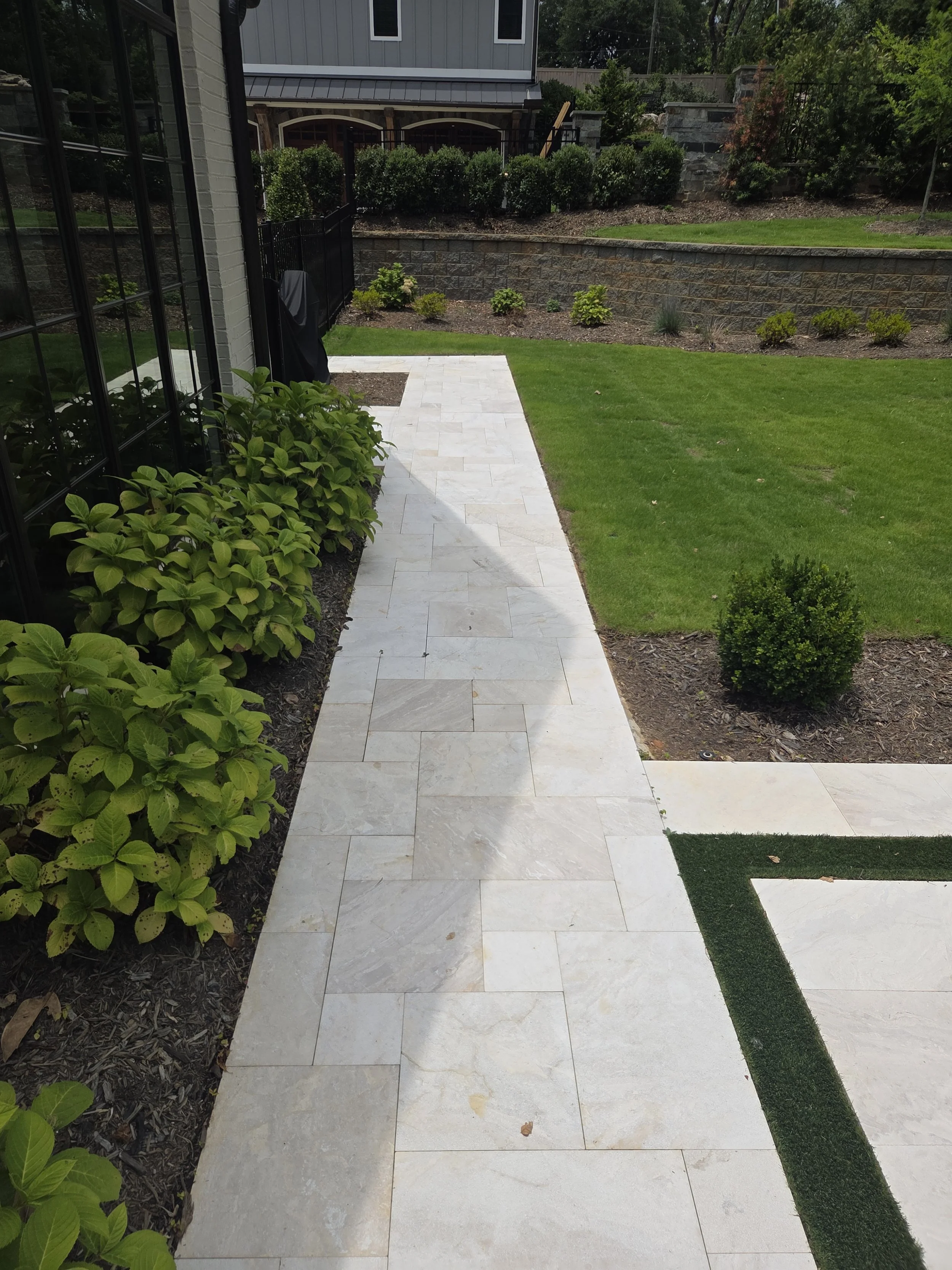 A backyard with a tiled walkway, green grass, and neatly trimmed bushes. A stone retaining wall separates the yard from an upper level with more bushes and trees. A house with gray siding is visible in the background.